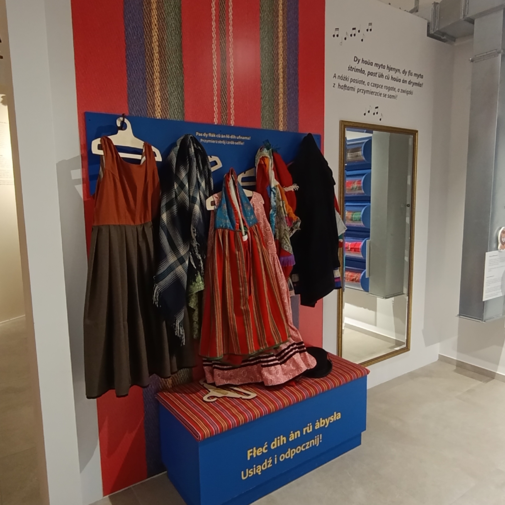 "Wnętrze muzeum z ekspozycją tradycyjnych strojów regionalnych zawieszonych na wieszakach na tle kolorowej ściany. Po prawej stronie lustro i rolki tkanin w kratę zamontowane na ścianie. W tle widoczna sylwetka postaci w stroju duchownego oraz obrazy i de