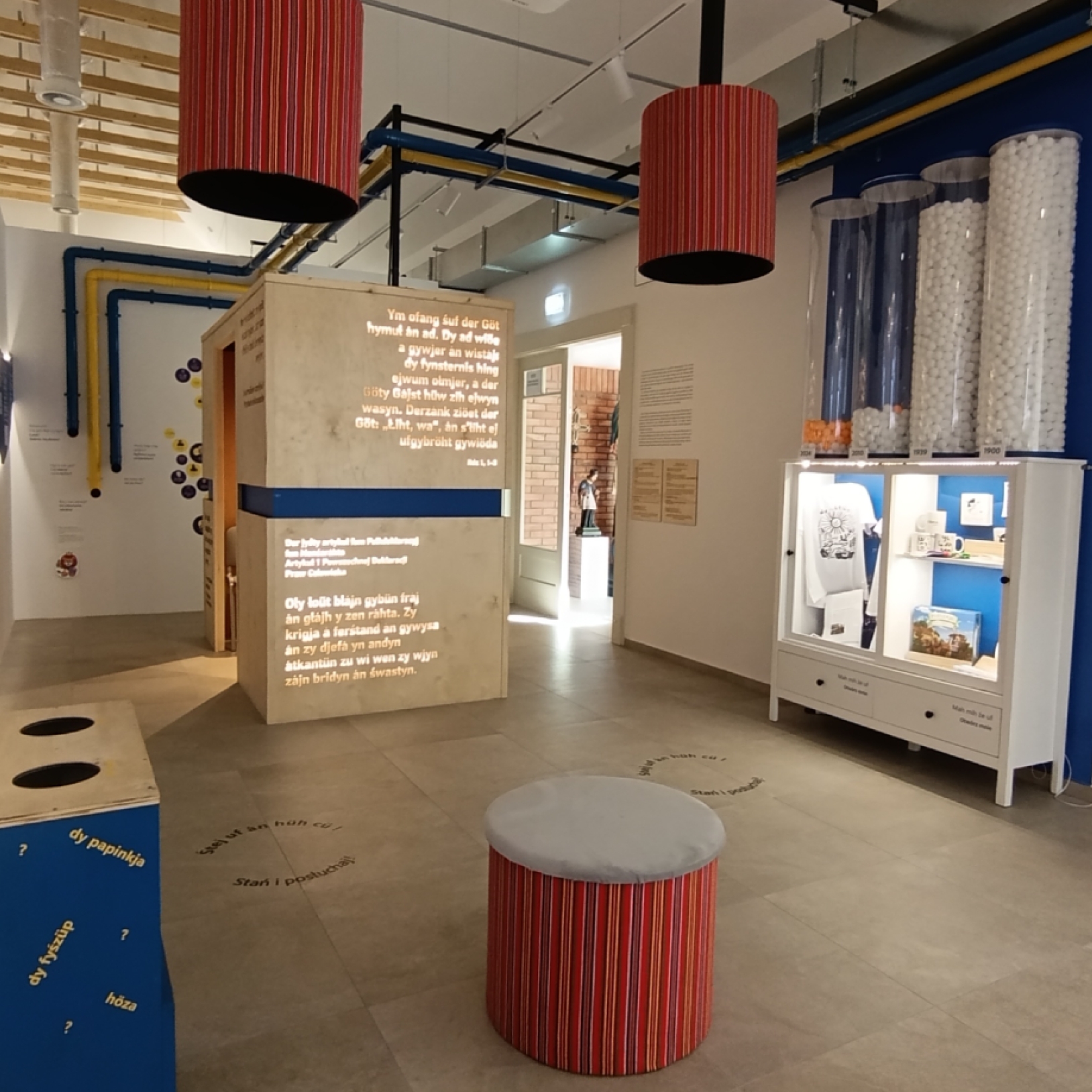 "Nowoczesna sala muzealna z interaktywną ekspozycją. Po lewej stronie ściana z niebieską linią czasową i fotografiami, po prawej instalacja z rurami i przezroczystymi tubami z piłeczkami. W centralnej części sześcienna konstrukcja z tekstami oraz gablotka