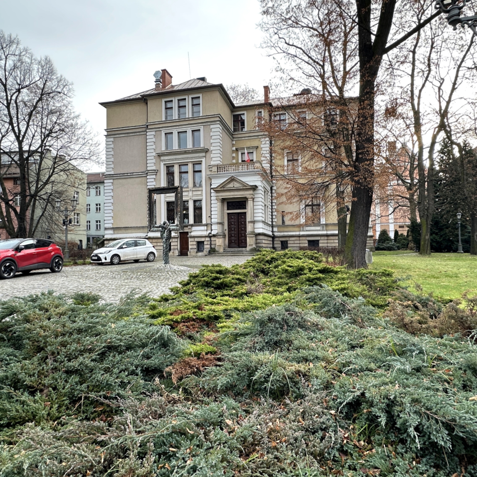 "Willa Caro w Gliwicach widziana zza kutego ogrodzenia z czerwoną ceglaną podmurówką. Na pierwszym planie znajduje się rzeźba leżącego lwa z czarnego materiału. Budynek ma klasycystyczną architekturę, jasną elewację i ozdobne gzymsy."