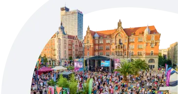 Fotografia tłumu ludzi zgromadzonego na miejskim placu przed sceną koncertową. W tle widać duży, zabytkowy budynek z czerwonej cegły oraz wysoki, nowoczesny, szklany wieżowiec. Zdjęcie ma zaokrąglone wycięcie w lewej górnej części, z jasnym tłem.