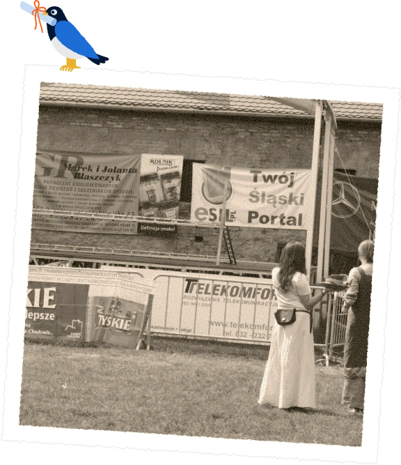 Czarno-białe zdjęcie plenerowego wydarzenia z banerami reklamowymi, w tym 'Twój Śląski Portal'. Na pierwszym planie dwie kobiety stojące na trawie.