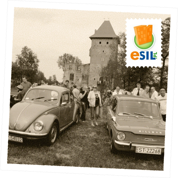 Czarno-białe zdjęcie plenerowego zlotu miłośników zabytkowych aut. Na pierwszym planie dwa klasyczne samochody – Volkswagen Garbus i Hillman, a w tle ruiny średniowiecznego zamku i zgromadzeni ludzie.