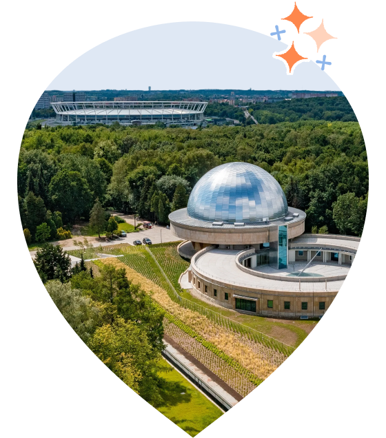 Zdjęcie Planetarium Śląskiego w Chorzowie otoczonego zielenią. W tle widoczny Stadion Śląski. W górnym rogu znajduje się grafika błyszczących gwiazdek.