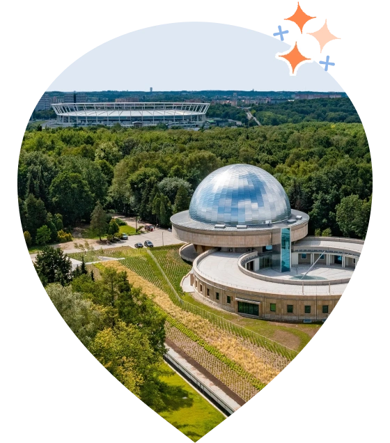 Zdjęcie Planetarium Śląskiego w Chorzowie otoczonego zielenią. W tle widoczny Stadion Śląski. W górnym rogu znajduje się grafika błyszczących gwiazdek.