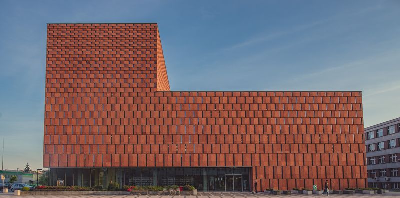 Fasada budynku biblioteki CINiBA z czerwonej okładziny, o geometrycznym wzorze. Na dole widać wejście z przeszklonymi drzwiami, przed którym znajdują się donice z roślinnością i ławki.