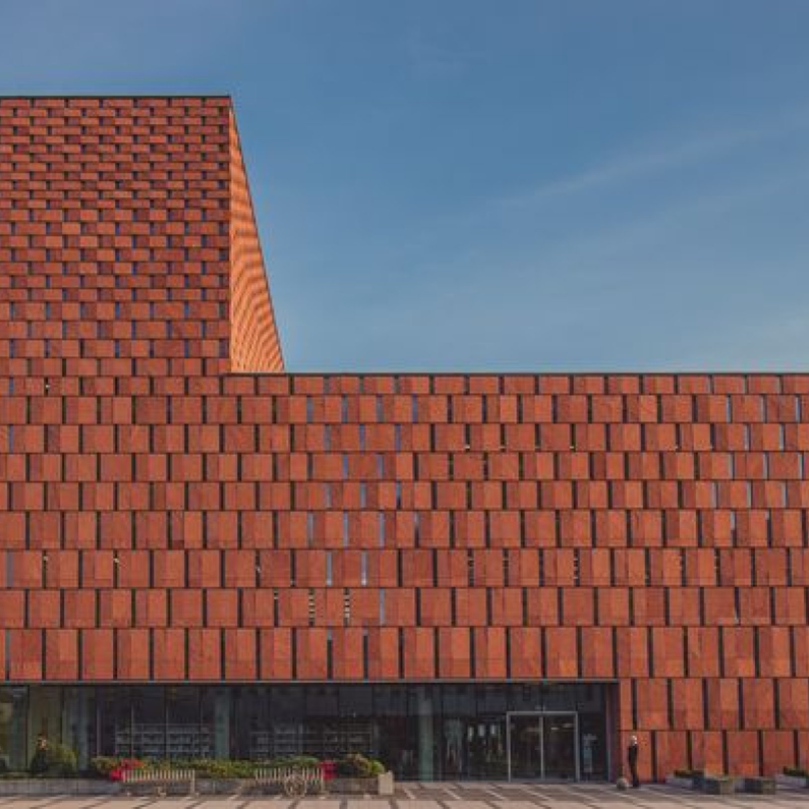 Fasada budynku biblioteki CINiBA z czerwonej okładziny, o geometrycznym wzorze. Na dole widać wejście z przeszklonymi drzwiami, przed którym znajdują się donice z roślinnością i ławki.