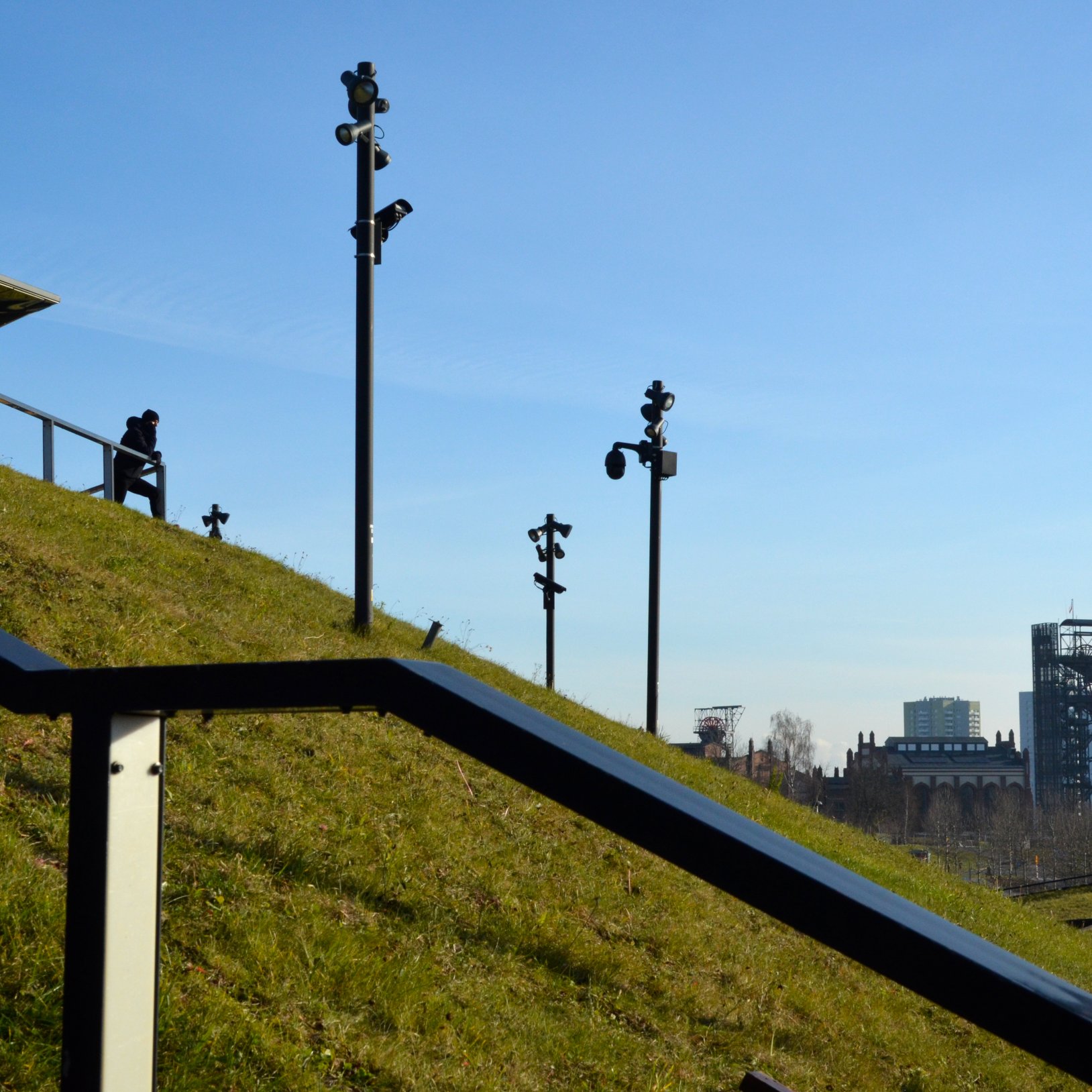 "Wzniesienie porośnięte trawą z zamontowanymi latarniami i kamerami monitoringu, tworzące punkt widokowy. Na górze osoba opiera się o barierkę, patrząc w dal. W tle widoczna panorama miasta z charakterystycznymi budynkami i błękitne niebo bez chmur."   },