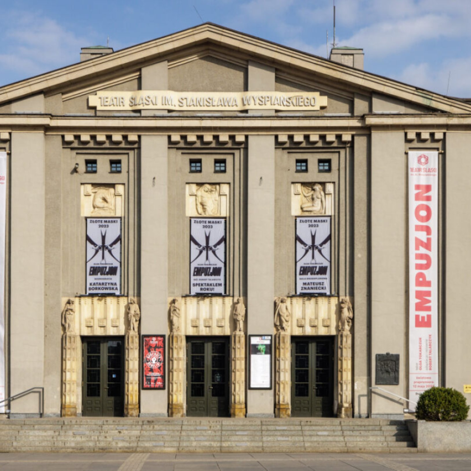 "Zbliżenie na front Teatru Śląskiego z trzema głównymi wejściami, ozdobionymi złotymi detalami i banerami spektaklu 'Empuzjon'. Widoczna także tablica z repertuarem."