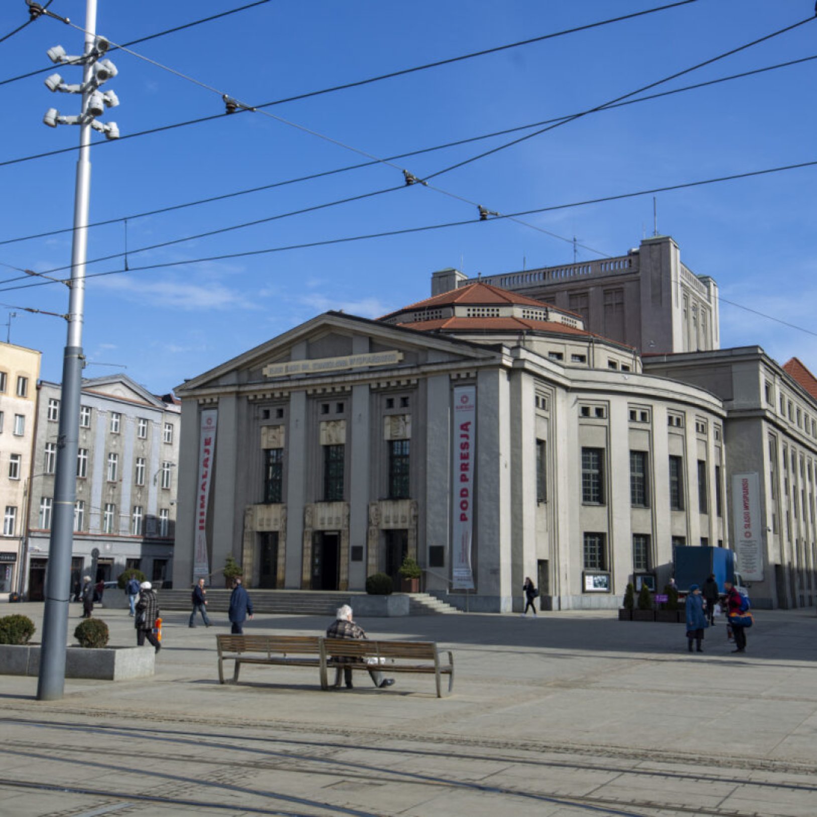 "Boczny widok na Teatr Śląski z napisem 'POD PRESJĄ' na banerze oraz dużym placem przed wejściem. W tle widać niebieskie niebo i kilka przechodzących osób."