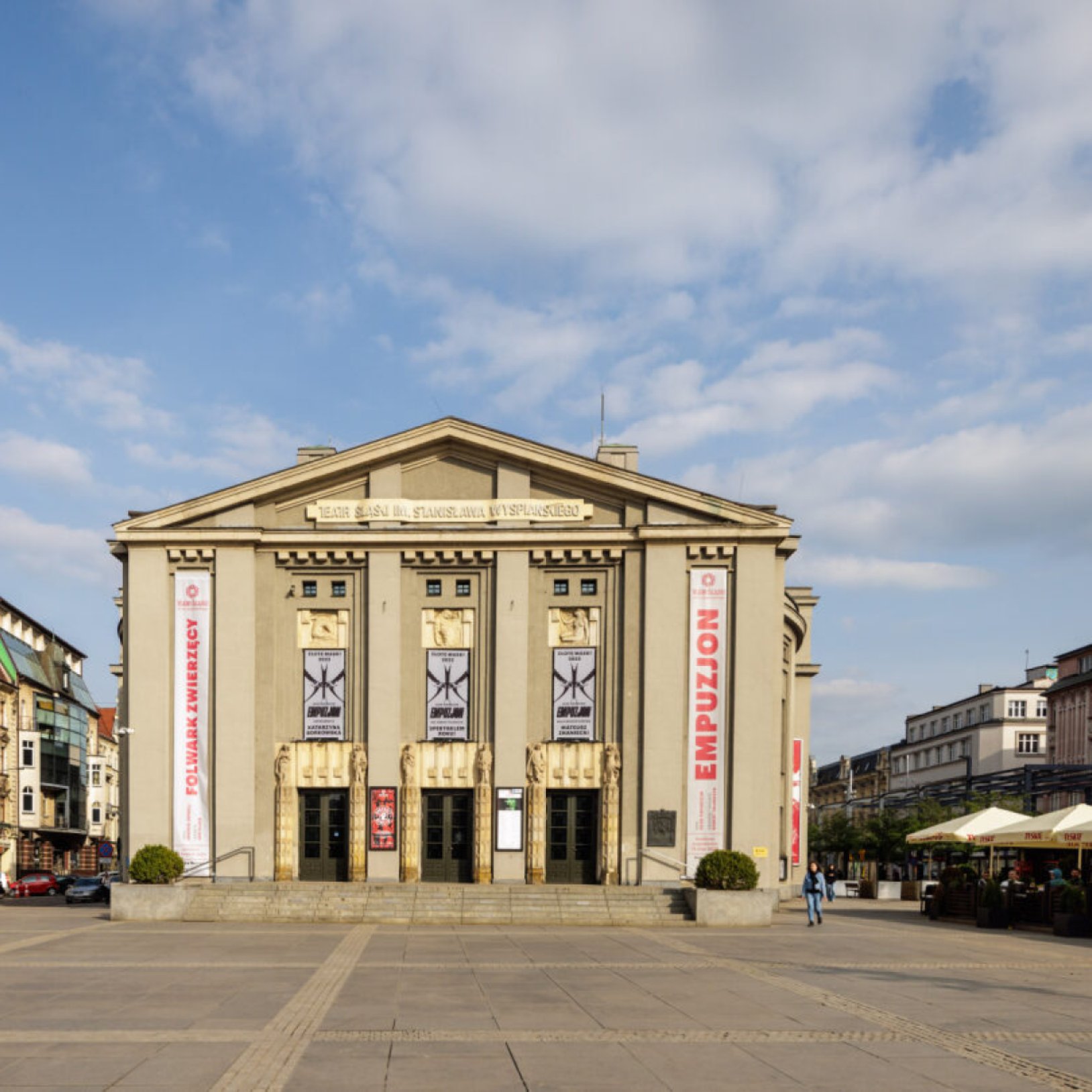 "Widok frontalny na Teatr Śląski im. Stanisława Wyspiańskiego z trzema wejściami, kolumnami oraz złotymi płaskorzeźbami nad drzwiami. Po bokach wiszą banery promujące spektakle."
