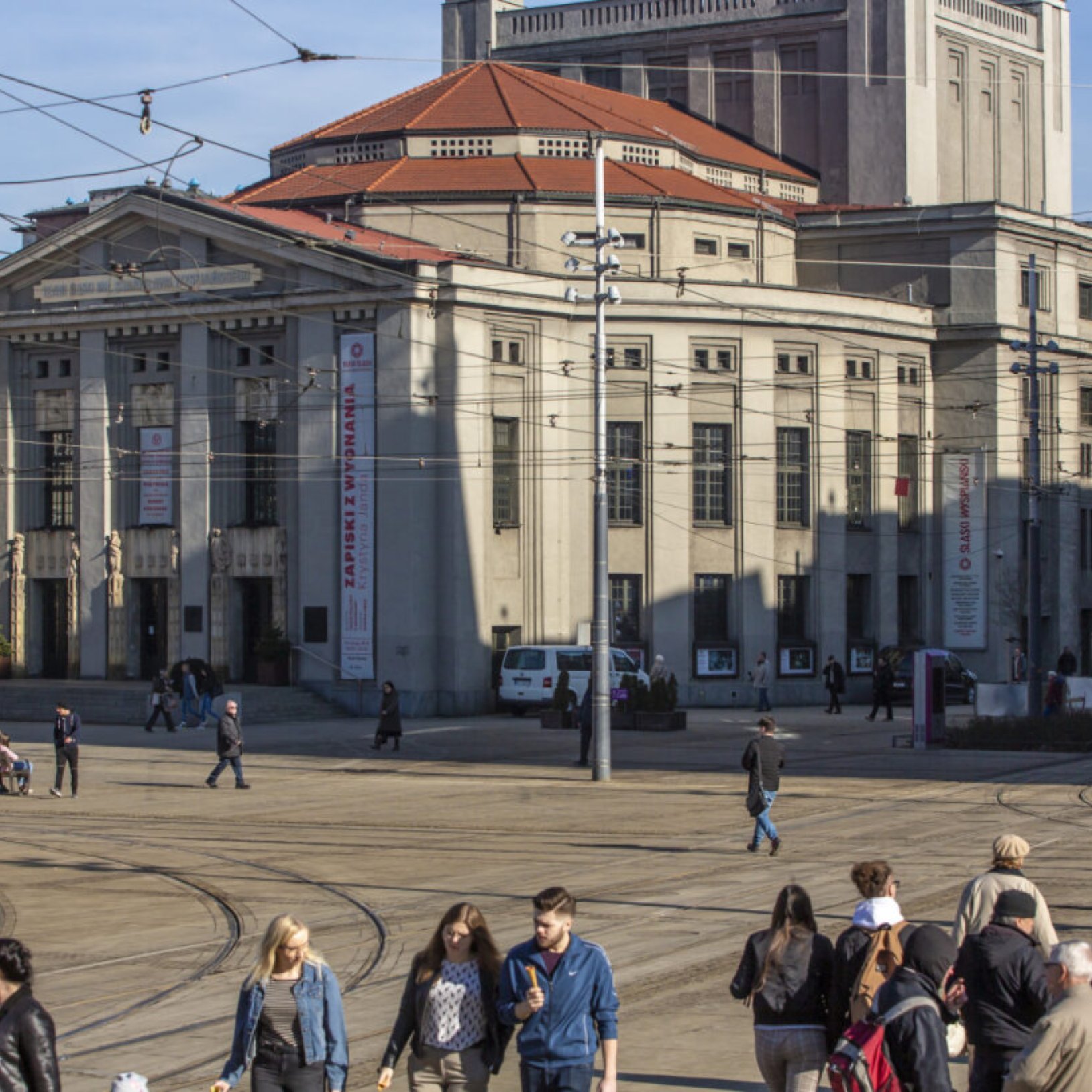 "Teatr Śląski z widokiem na boczną i frontową elewację. Na placu przed budynkiem widoczne są osoby spacerujące oraz tramwajowe przewody trakcyjne."