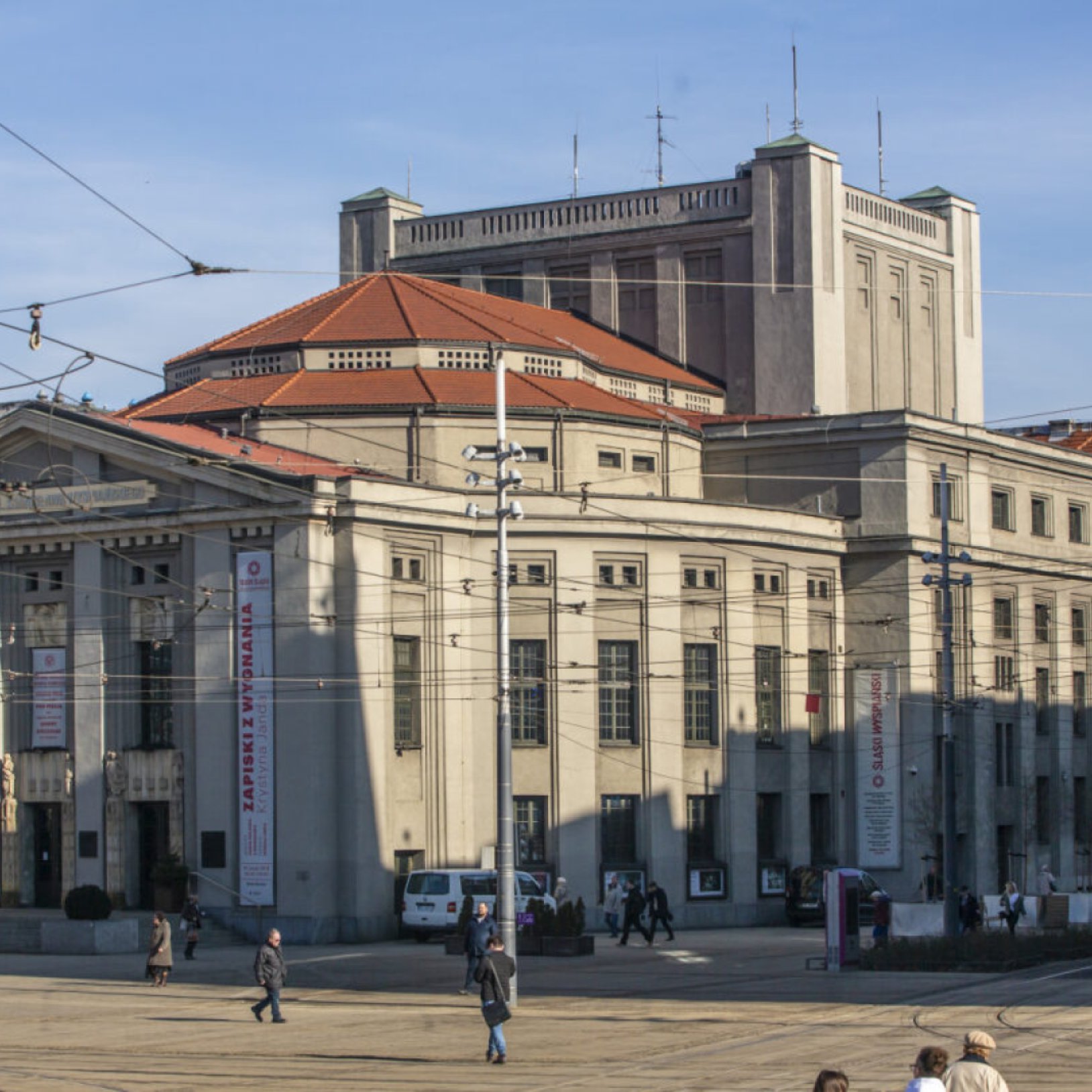 "Zabytkowy budynek Teatru Śląskiego z kolumnowym wejściem, czerwonym dachem i wieżą w tle. Przed gmachem widać plac z pieszymi oraz tory tramwajowe."