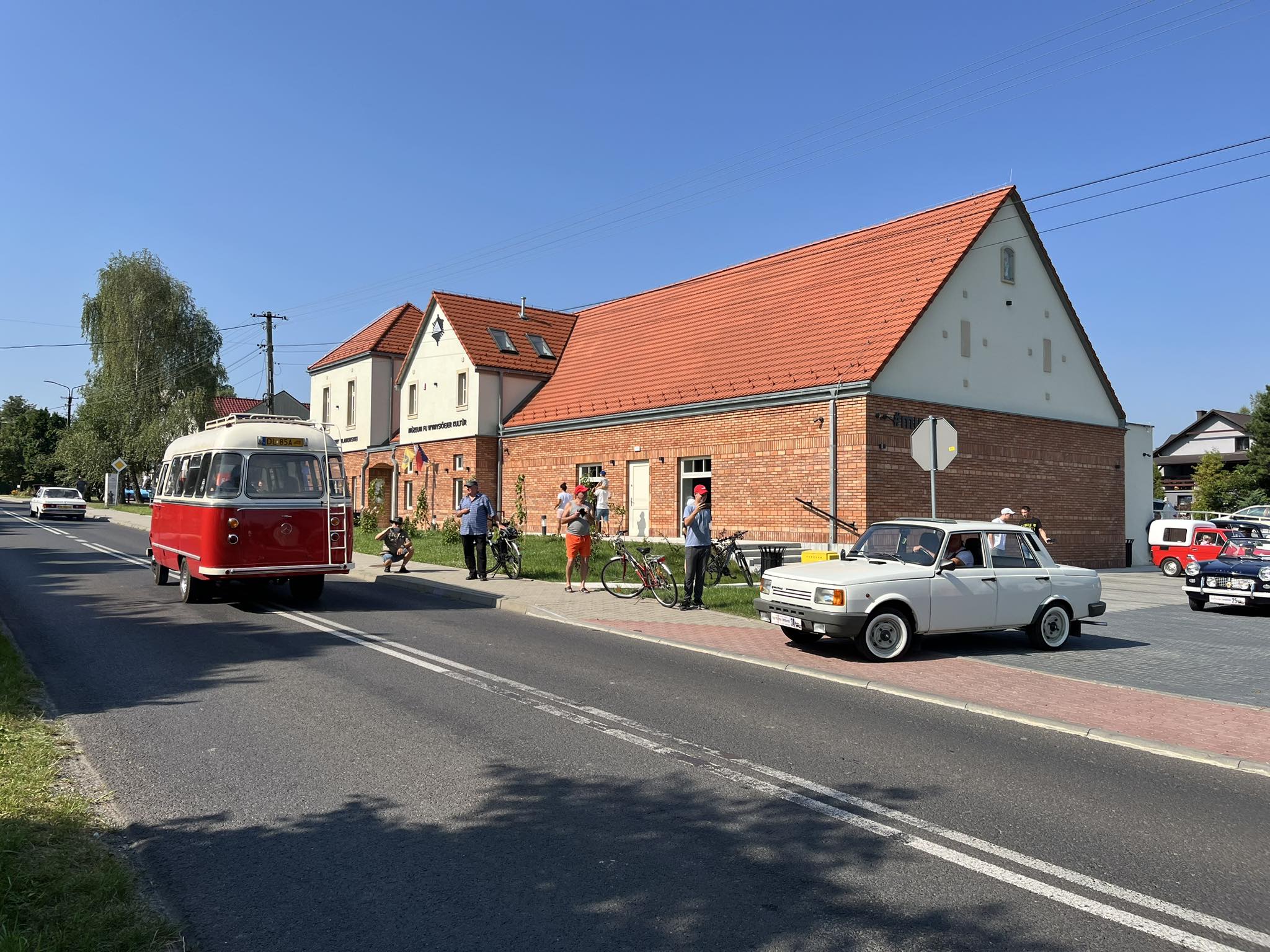 "Zewnętrzny widok na budynek z czerwonej cegły z białymi elementami elewacji i czerwonym dachem. Przed budynkiem zaparkowany zabytkowy czerwono-biały autobus oraz biały samochód osobowy. Wokół znajdują się ludzie z rowerami, a w tle niebieskie niebo i nap