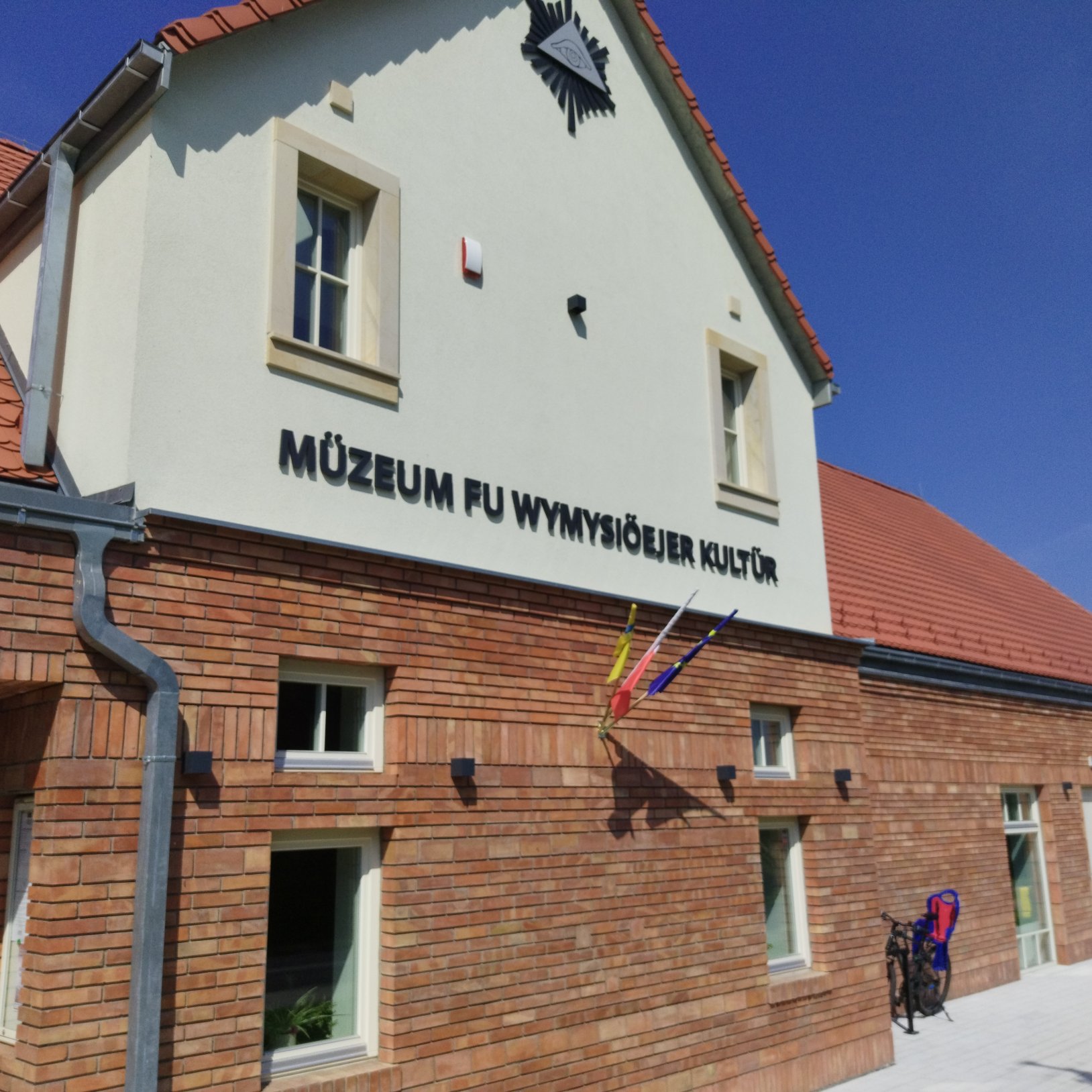 "Zbliżenie na fasadę budynku z czerwonej cegły z napisem 'Müzeum fu Wymysiöejer Kultur' oraz trzema kolorowymi flagami. Nad wejściem symbol w kształcie oka w promieniach. Widoczna część dachu pokrytego czerwoną dachówką, a przy ścianie stojące rowery."