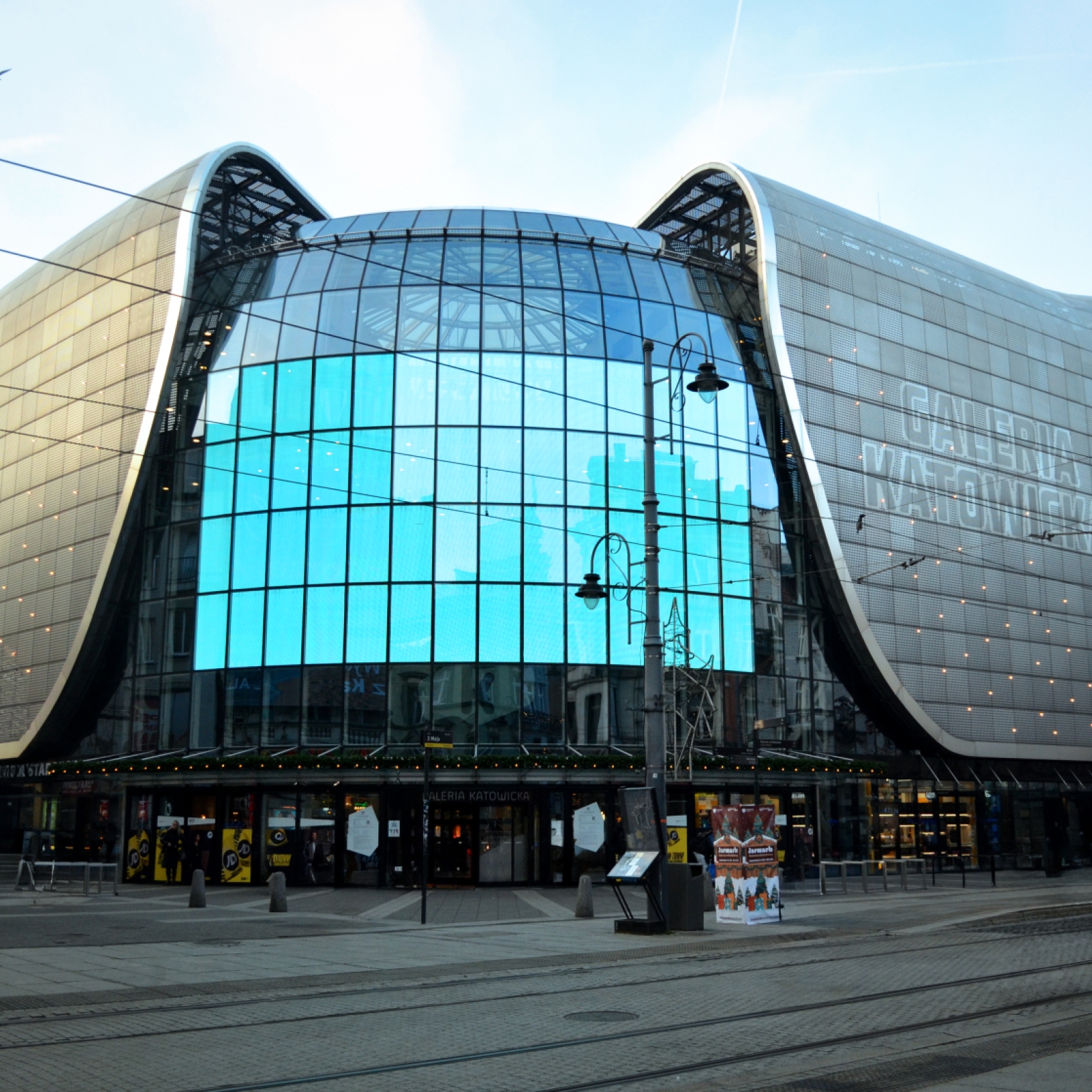 "Nowoczesna fasada Galerii Katowickiej z przeszklonym, zakrzywionym frontem i dużym ekranem LED. Budynek ma zaokrąglone, metaliczne skrzydła po bokach. Na dole widoczne są wejścia oraz witryny sklepowe."