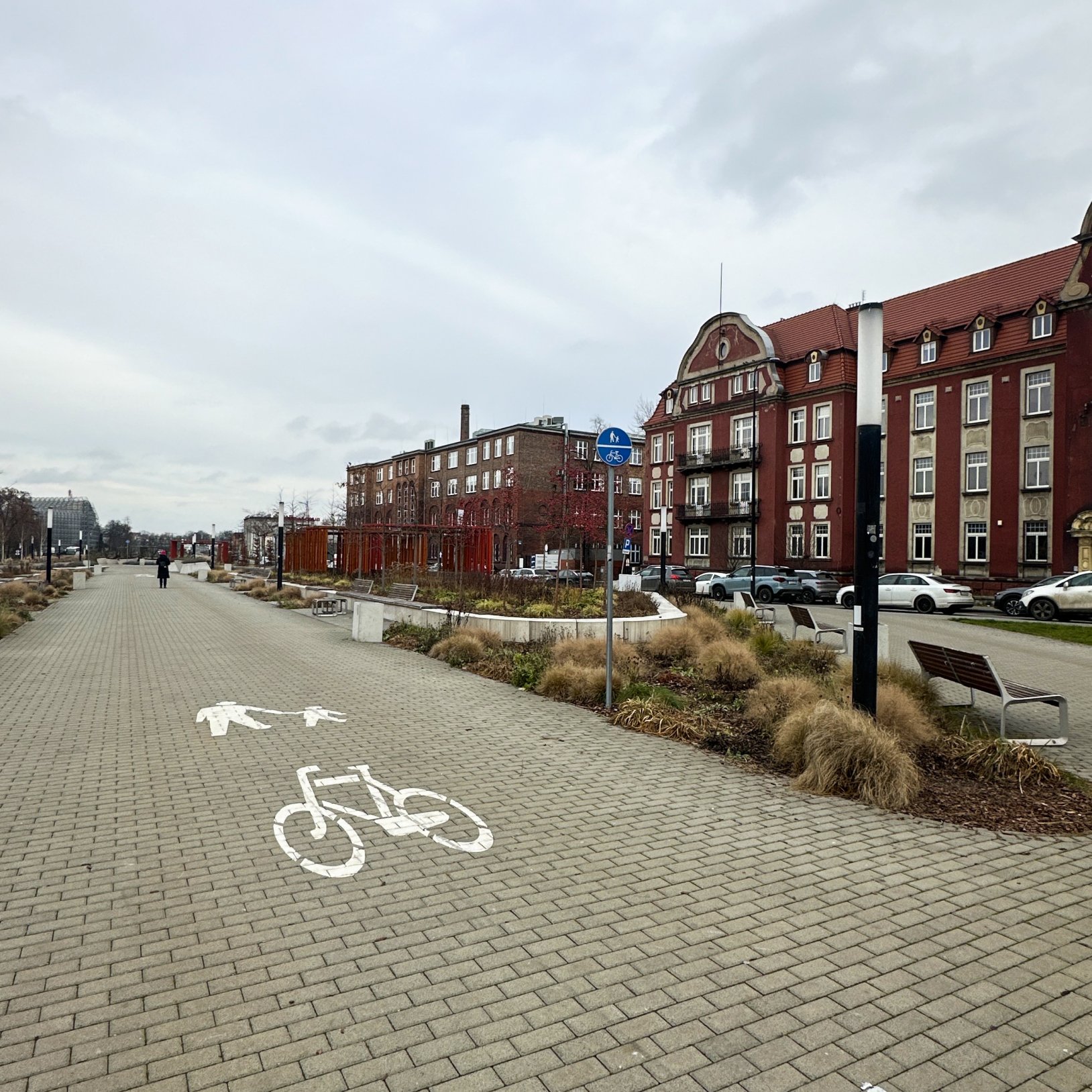 "Szeroki chodnik z wyznaczoną ścieżką pieszo-rowerową, oznaczoną białymi symbolami na kostce brukowej. Po prawej stronie znajduje się rząd ceglanych kamienic z czerwonymi dachami. Wzdłuż ścieżki rosną ozdobne trawy i krzewy. Na końcu ścieżki widać sylwetk