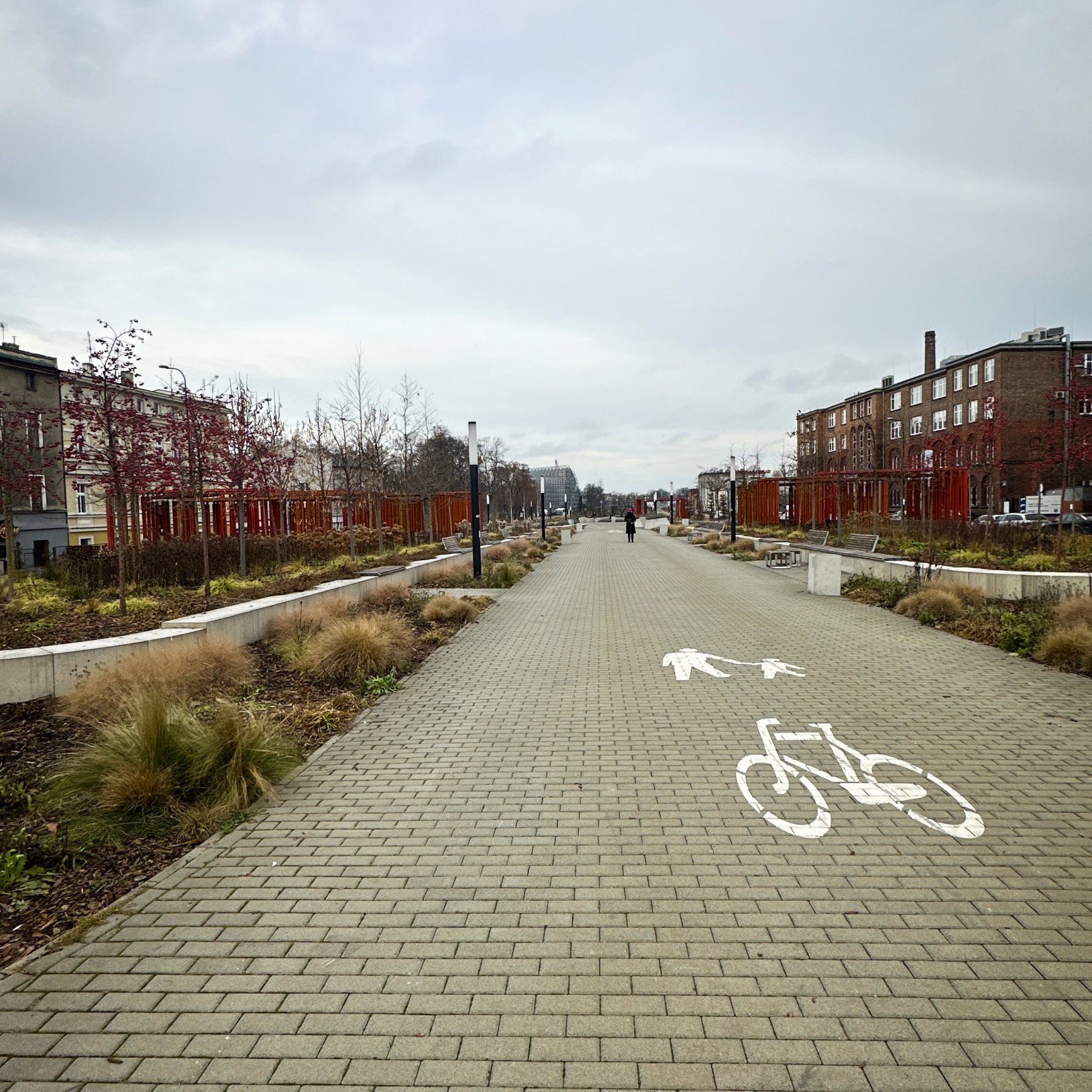 "Perspektywiczny widok wzdłuż szerokiej brukowanej alei z oznaczeniem dla pieszych i rowerzystów. Po obu stronach ścieżki znajdują się rabaty z ozdobnymi trawami i młodymi drzewami, otoczone betonowymi obrzeżami. W oddali widać spacerujących ludzi i dalsz