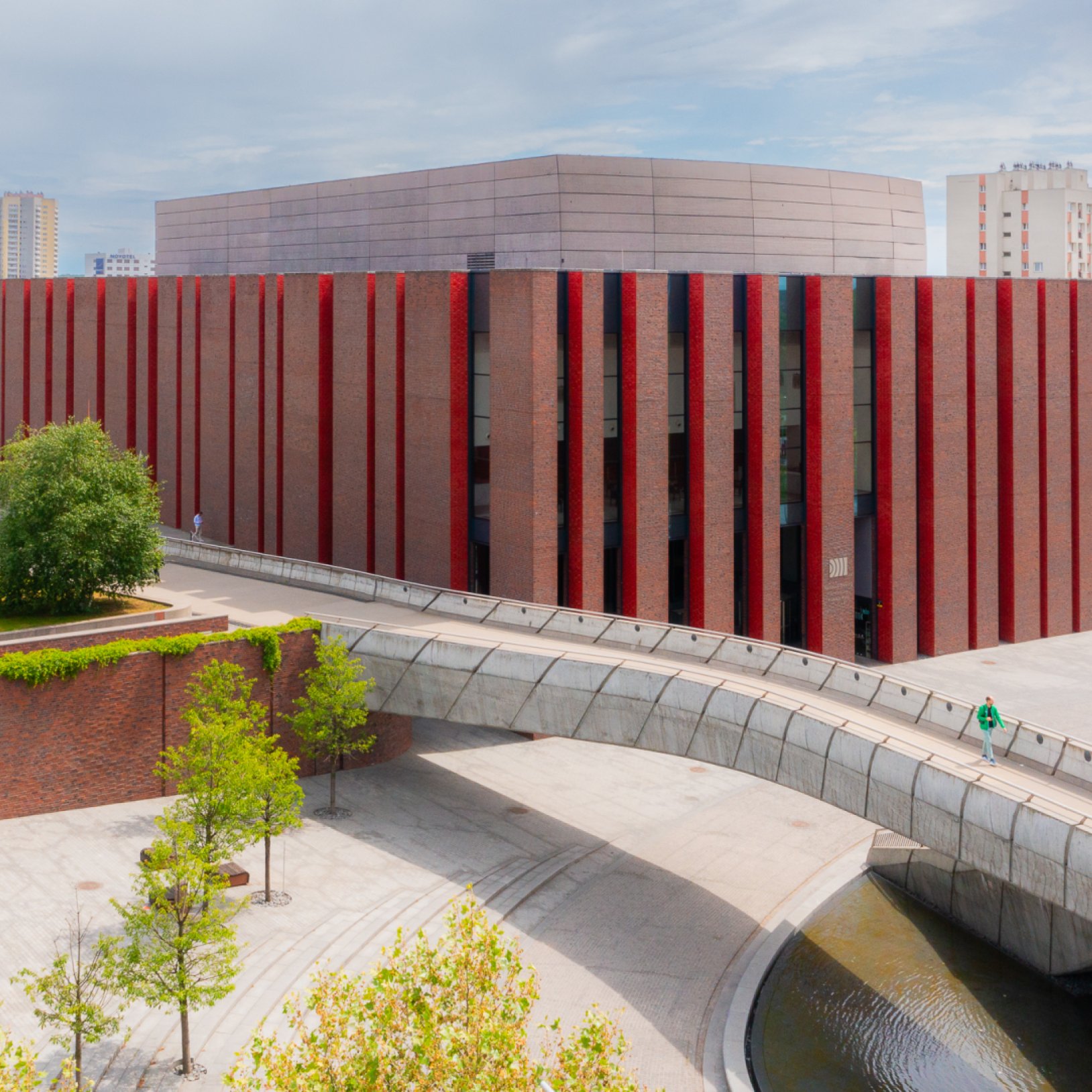 "Zbliżenie na elewację budynku NOSPR w Katowicach z czerwono-czarnymi pionowymi pasami i przeszklonymi wejściami. Na pierwszym planie biegnie zakrzywiona kładka nad kanałem wodnym."