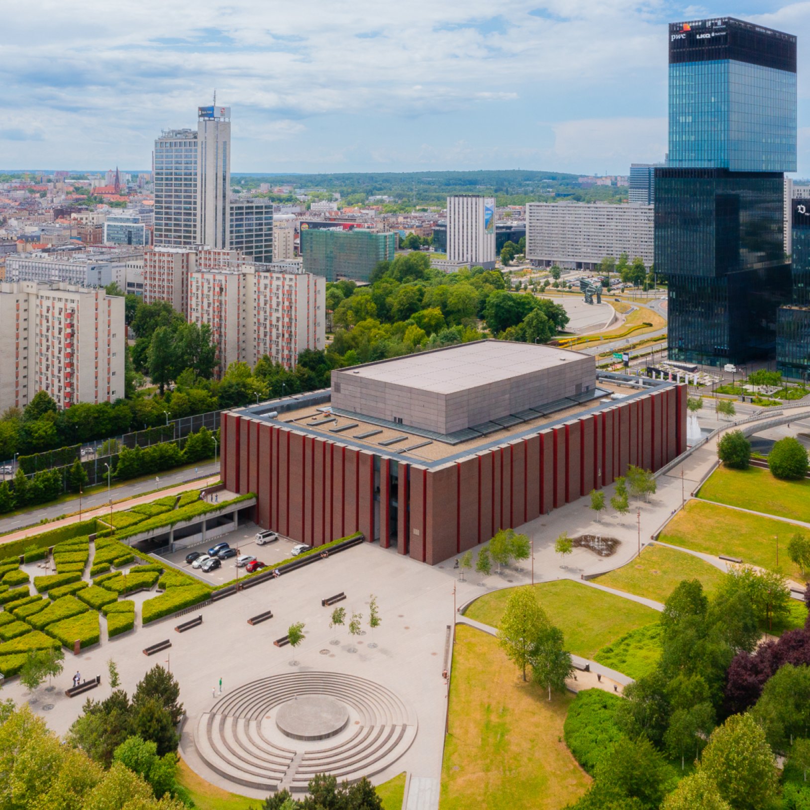 "Widok z lotu ptaka na nowoczesny budynek Narodowej Orkiestry Symfonicznej Polskiego Radia w Katowicach, otoczony zielenią i miejską zabudową. Na pierwszym planie widoczny plac z kolistymi schodami i alejkami parkowymi."