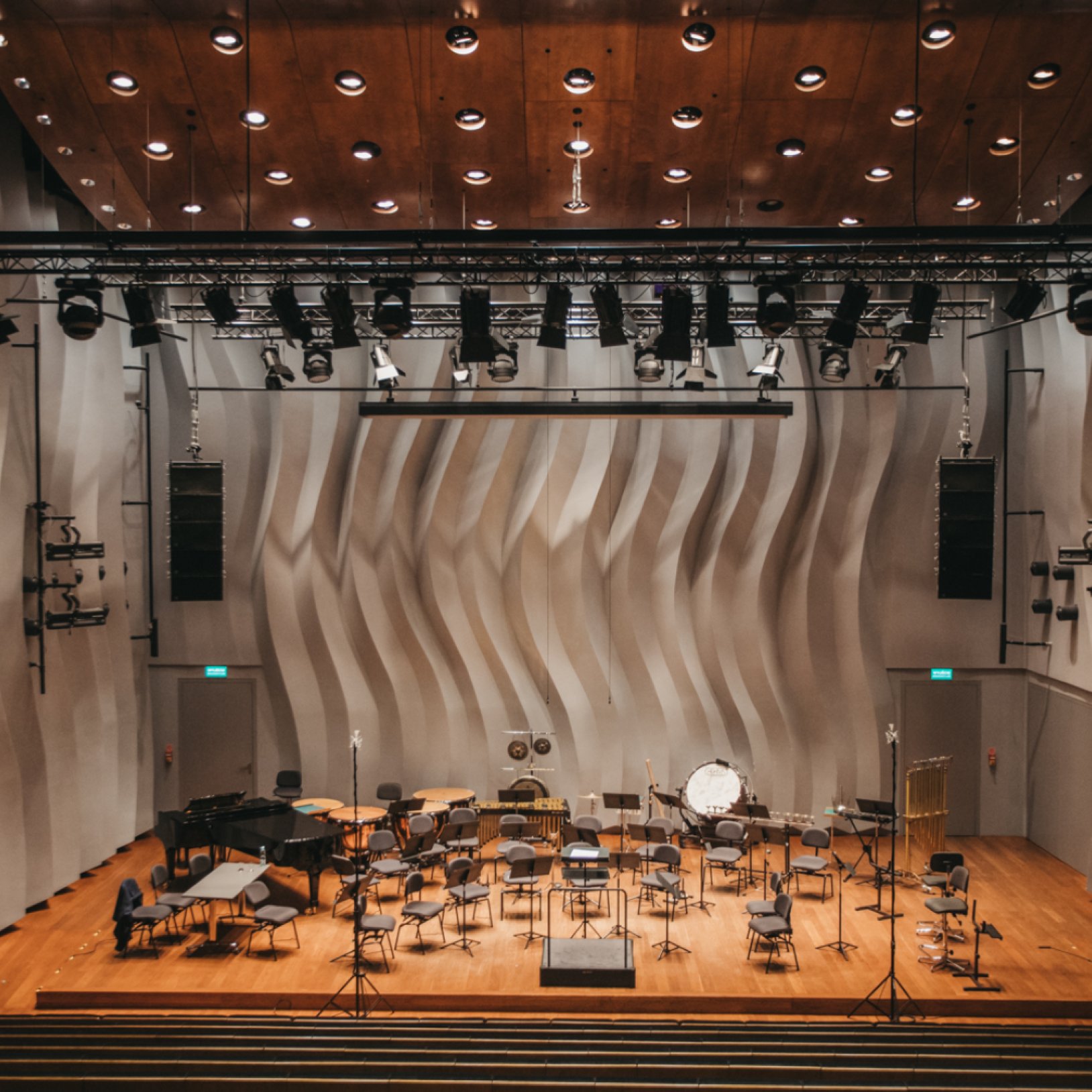 "Wnętrze nowoczesnej sali koncertowej z drewnianą sceną, licznymi krzesłami dla muzyków i instrumentami orkiestrowymi. W tle widoczna ściana z falistą fakturą oraz oświetlenie sufitowe i sprzęt nagłośnieniowy."