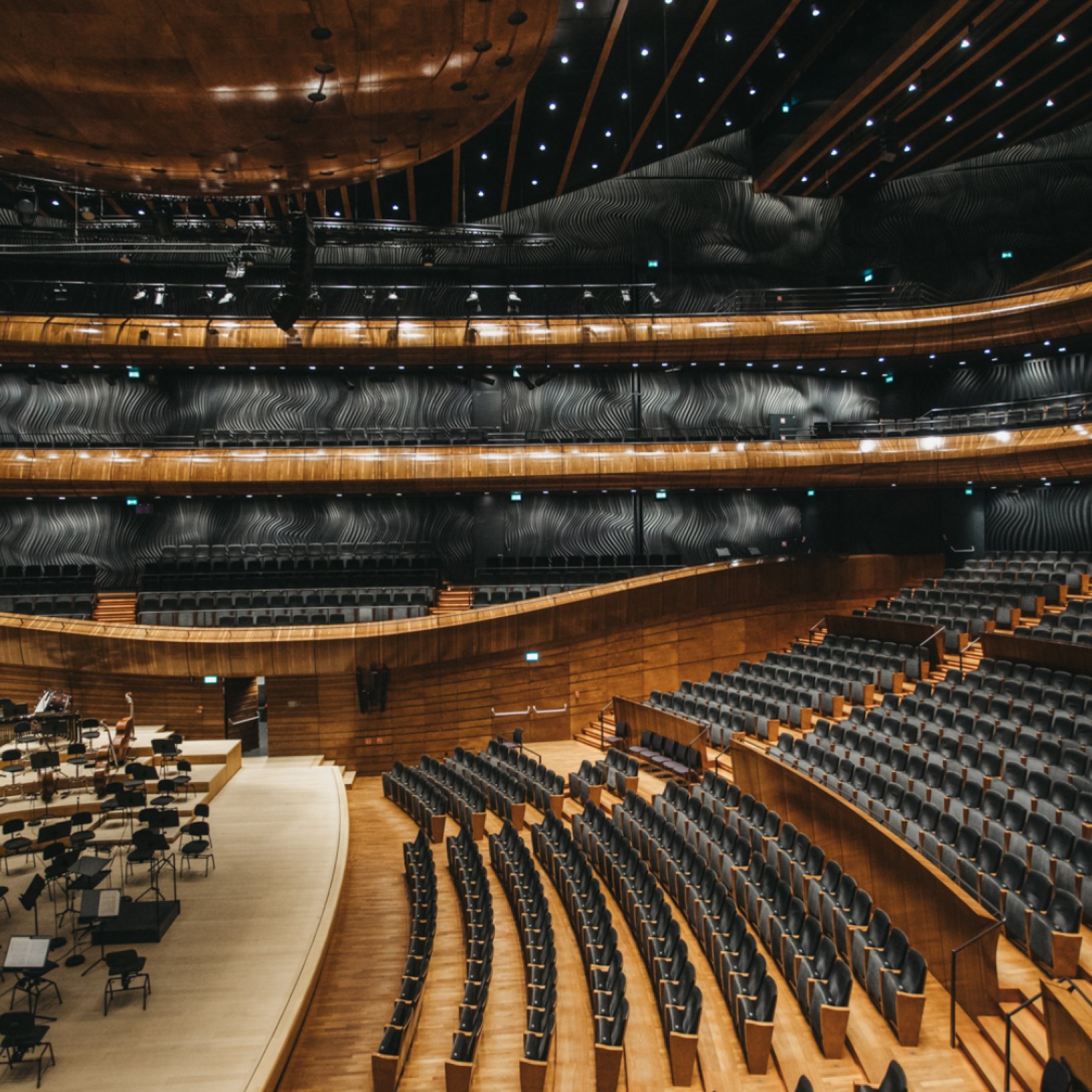 "Widok na wnętrze wielopoziomowej sali koncertowej z czarnymi fotelami, drewnianymi balustradami i sufitami ozdobionymi punktowym oświetleniem. Po lewej stronie widoczna część sceny z rozstawionymi pulpitami i krzesłami."