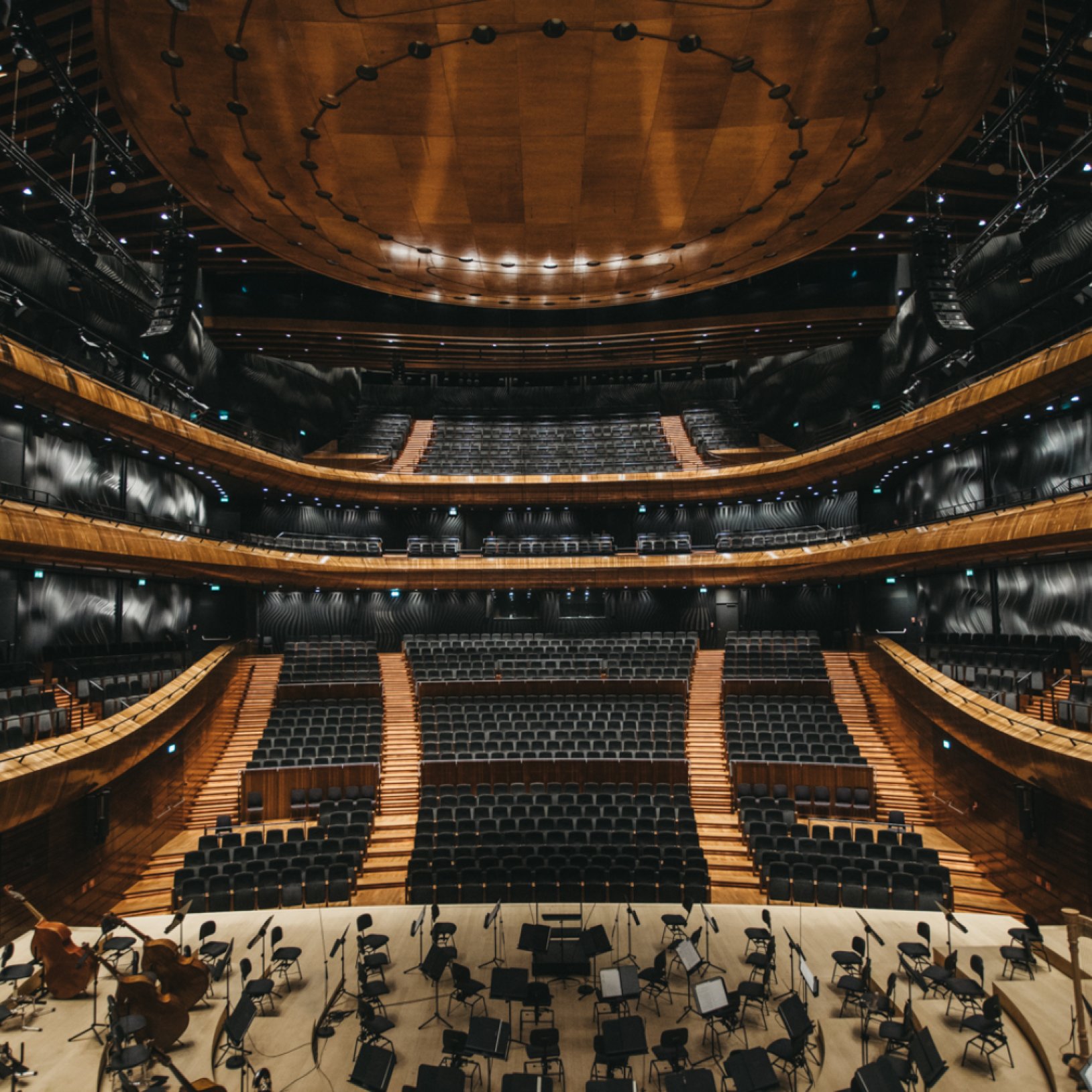 "Nowoczesna sala koncertowa z kilkoma poziomami balkonów, wykończona w ciemnym drewnie i czerni. Widok z perspektywy sceny, gdzie stoją pulpity i krzesła dla orkiestry."