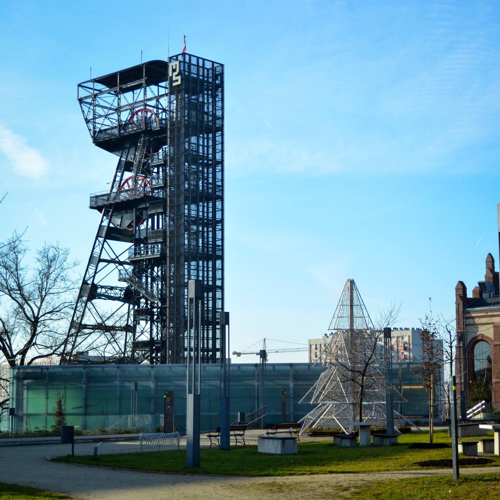 "Widok na teren Muzeum Śląskiego w Katowicach z wieżą wyciągową dawnej kopalni z numerem 'P2'. Obok znajdują się nowoczesne i zabytkowe budynki oraz metalowa konstrukcja w kształcie choinki. W tle widać fragmenty miasta i żurawie budowlane.