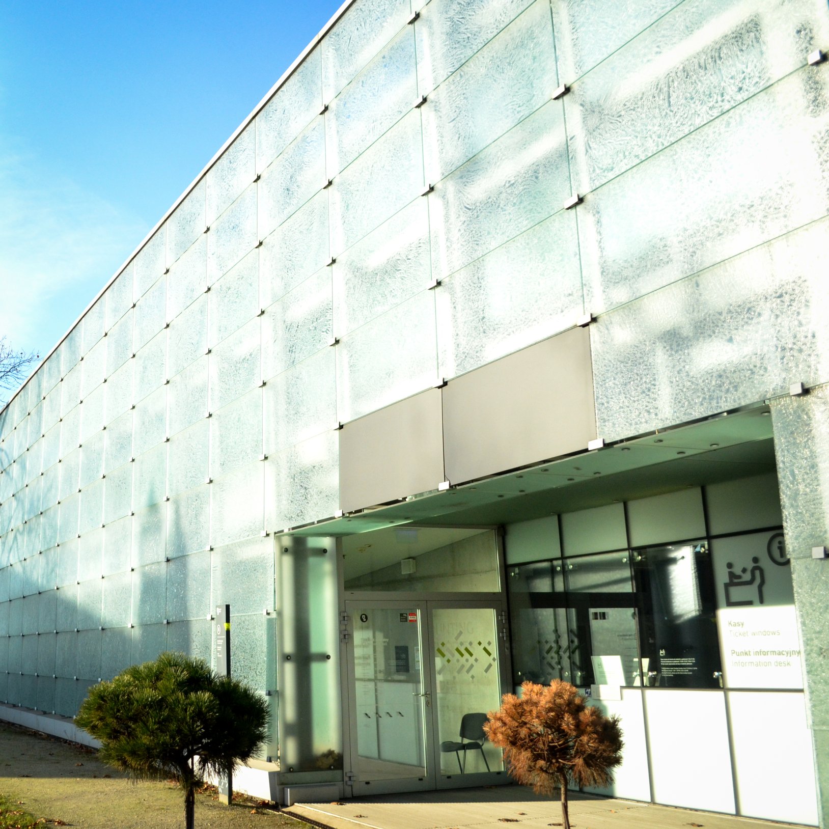 "Nowoczesny budynek z przeszkloną fasadą będący częścią Muzeum Śląskiego w Katowicach. Na zdjęciu widoczny jest główny punkt informacyjny i wejście do budynku. Wzdłuż ściany rosną małe drzewa ozdobne. Niebo jest jasne i bezchmurne."
