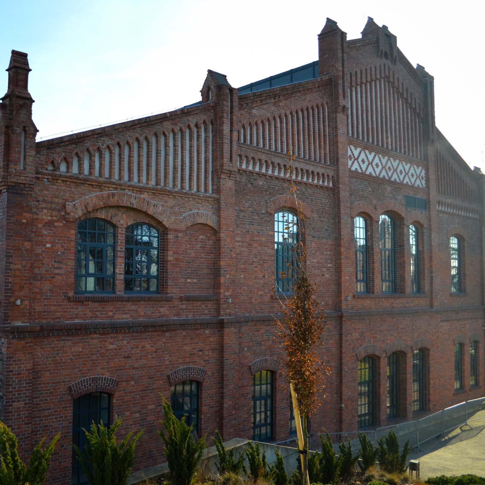 "Zabytkowy, ceglany budynek na terenie Muzeum Śląskiego w Katowicach. Fasada ozdobiona jest dekoracyjnym wzorem z cegieł i dużymi oknami łukowymi. Przed budynkiem rośnie kilka drzew i ozdobnych krzewów. Słoneczny dzień, błękitne niebo."