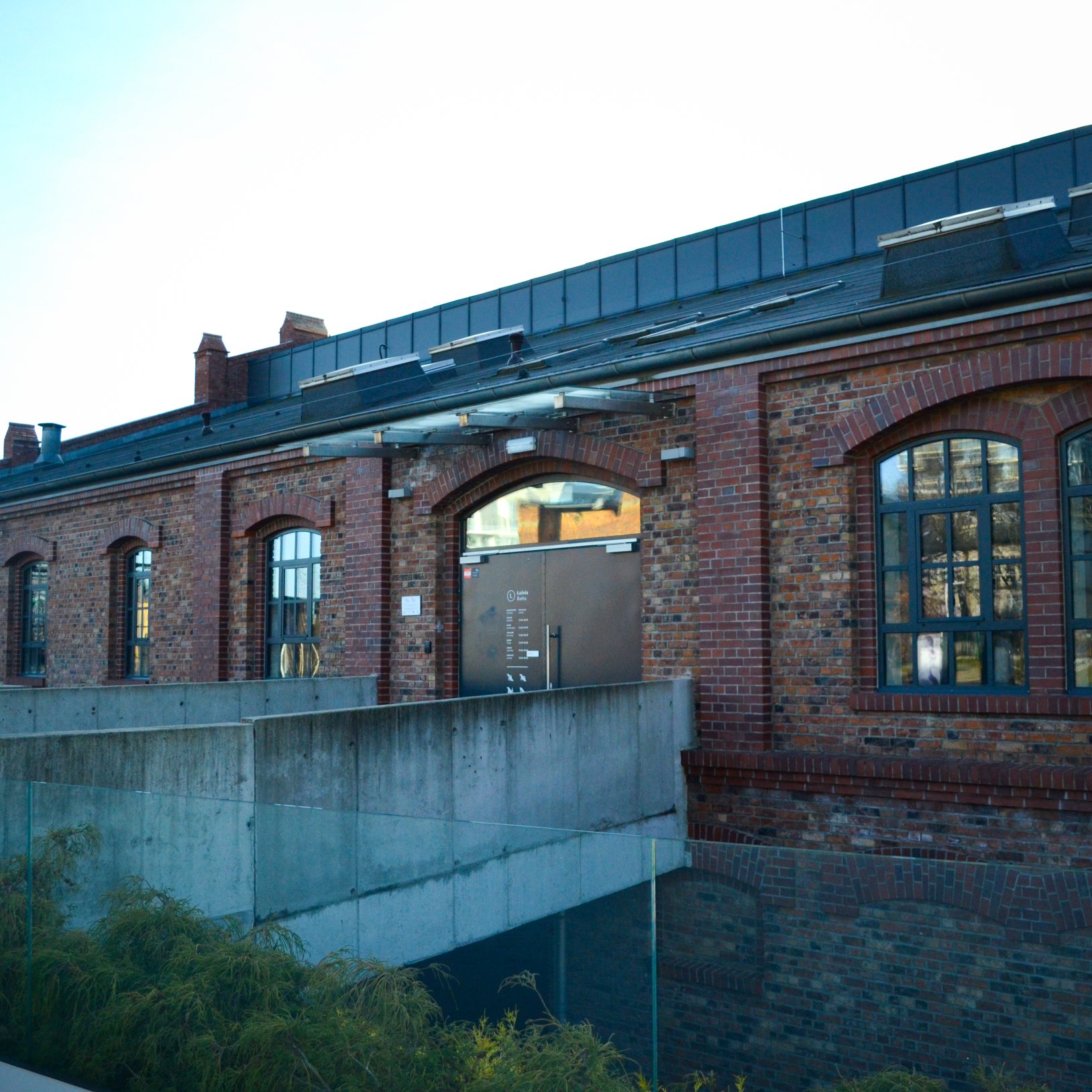 "Wejście do jednego z zabytkowych budynków Muzeum Śląskiego w Katowicach. Budynek z czerwonej cegły z dużymi oknami łukowymi, a do wejścia prowadzi betonowy podest z balustradą. Na dachu widoczne są nowoczesne elementy techniczne."