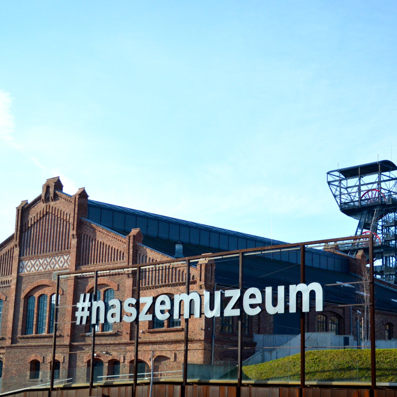 "Widok na ceglany budynek Muzeum Śląskiego z dużym napisem #naszemuzeum na przeszklonej konstrukcji w pierwszym planie. W tle widoczna jest wieża wyciągowa kopalni oraz nowoczesne elementy zabudowy. Niebo jasne i bezchmurne."