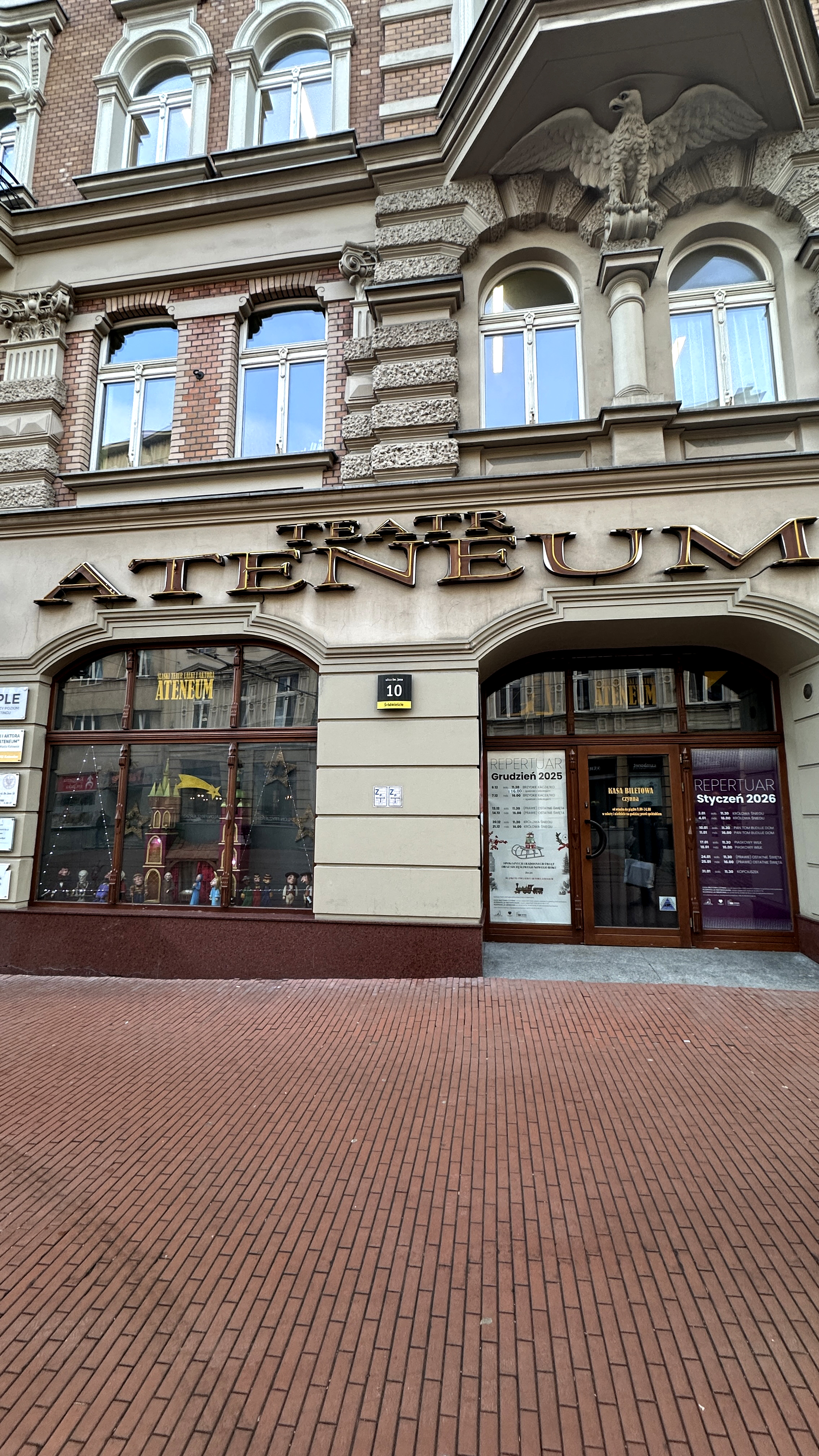 "Fasada Teatru Ateneum w Katowicach z ozdobną architekturą i dużym szyldem. Na witrynach widoczny jest repertuar na grudzień 2025 i styczeń 2026 oraz dekoracje teatralne w oknie wystawowym."