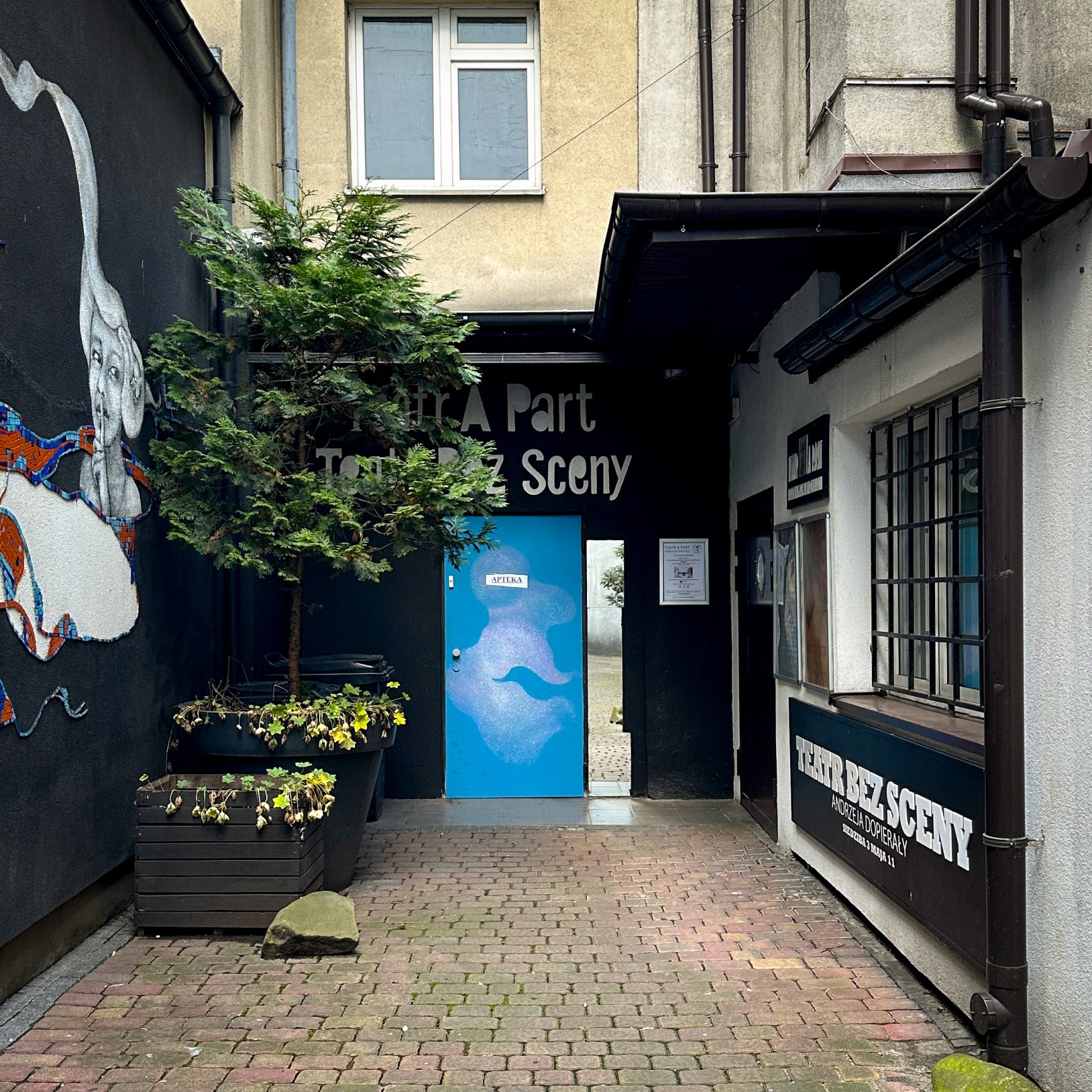"Podwórko z wejściem do teatru oznaczonego napisem 'Teatr Bez Sceny' i niebieskimi drzwiami. Po lewej stronie widoczny mural i donice z roślinami, po prawej okno z kratą i czarnym szyldem teatru."