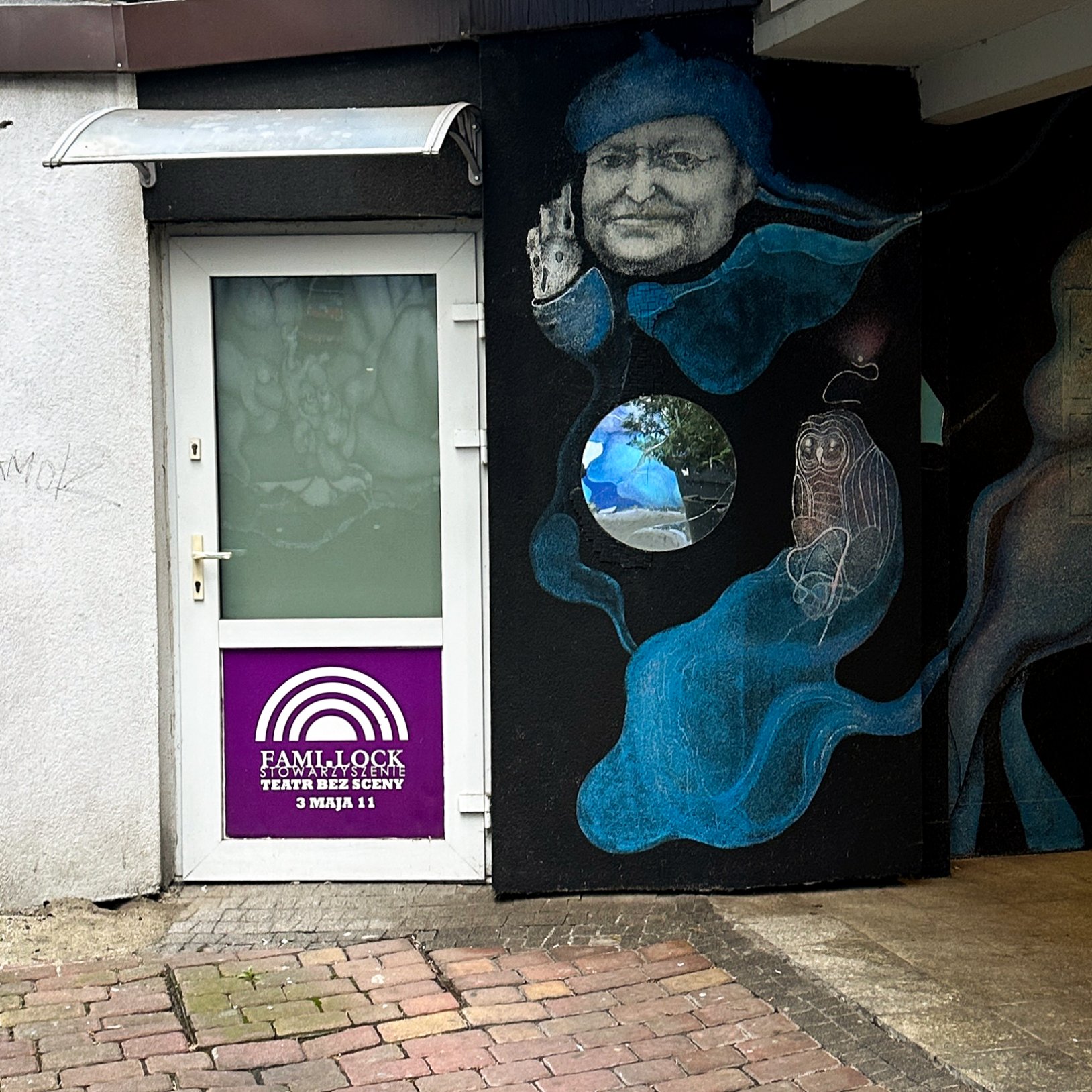 "Wejście do kameralnego teatru z białymi drzwiami i fioletowym logotypem 'FAMILOCK Stowarzyszenie Teatr Bez Sceny'. Obok znajduje się mural z twarzą mężczyzny oraz motywami niebieskiego dymu i lustra."