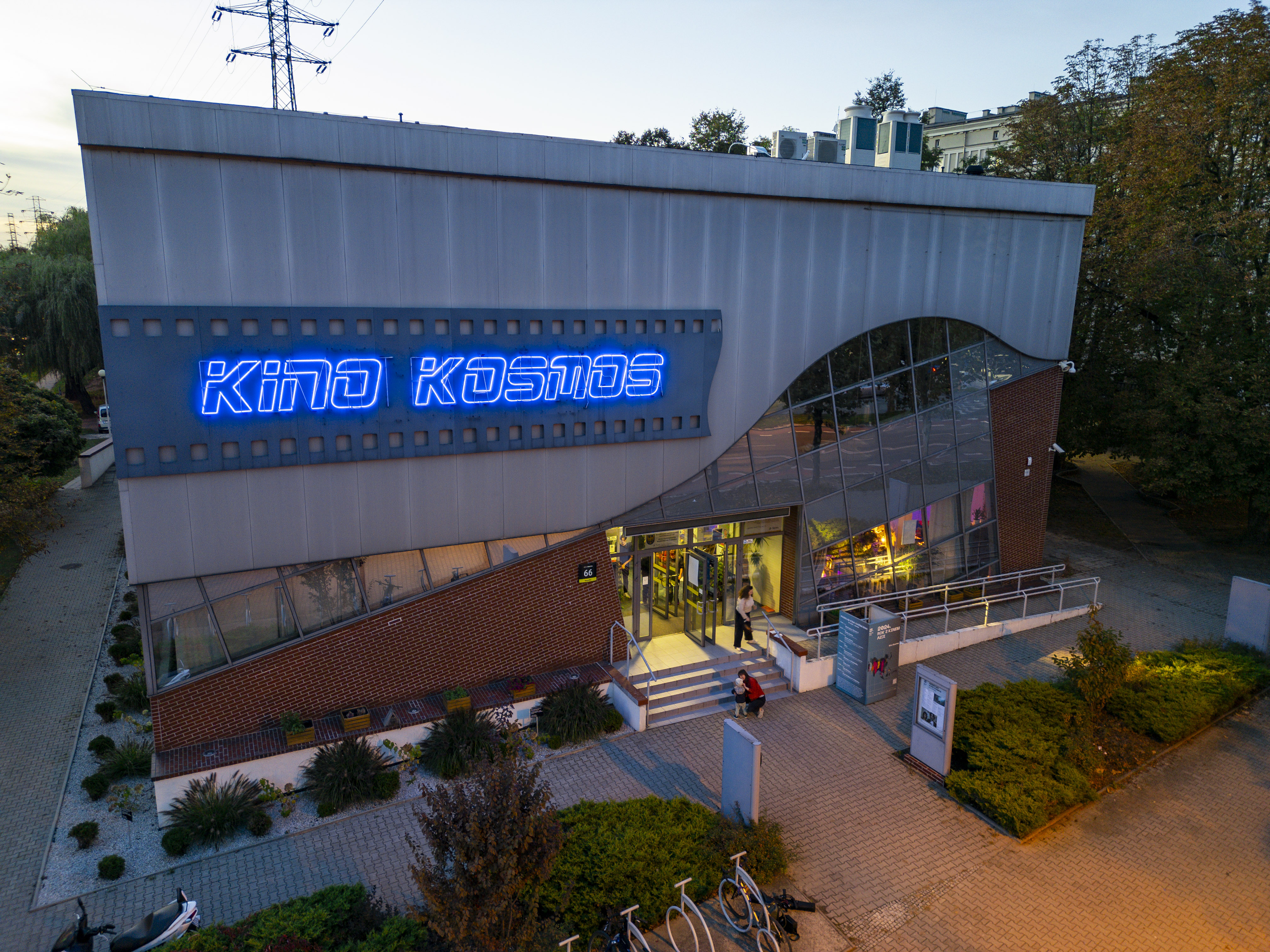 "Zewnętrzny widok na Kino Kosmos z charakterystycznym neonowym napisem w kolorze niebieskim. Budynek posiada przeszkloną fasadę i wejście z podświetlanymi schodami."