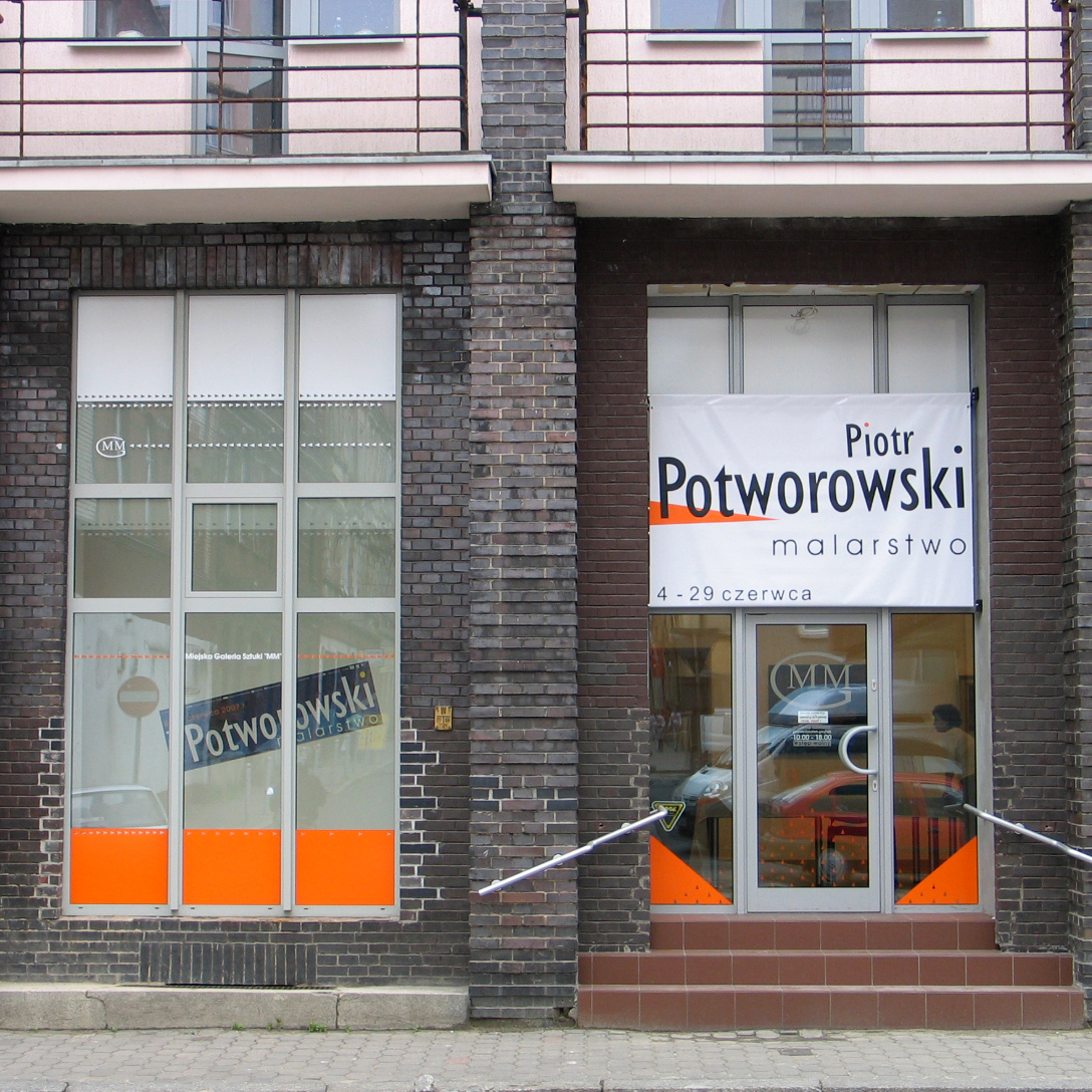 Wejście do galerii sztuki mieszczącej się w budynku z ciemnej cegły. Na drzwiach zawieszony baner wystawy 'Piotr Potworowski – malarstwo', z datami '4–29 czerwca'. Wystawa promowana jest także plakatem widocznym w oknie po lewej stronie.