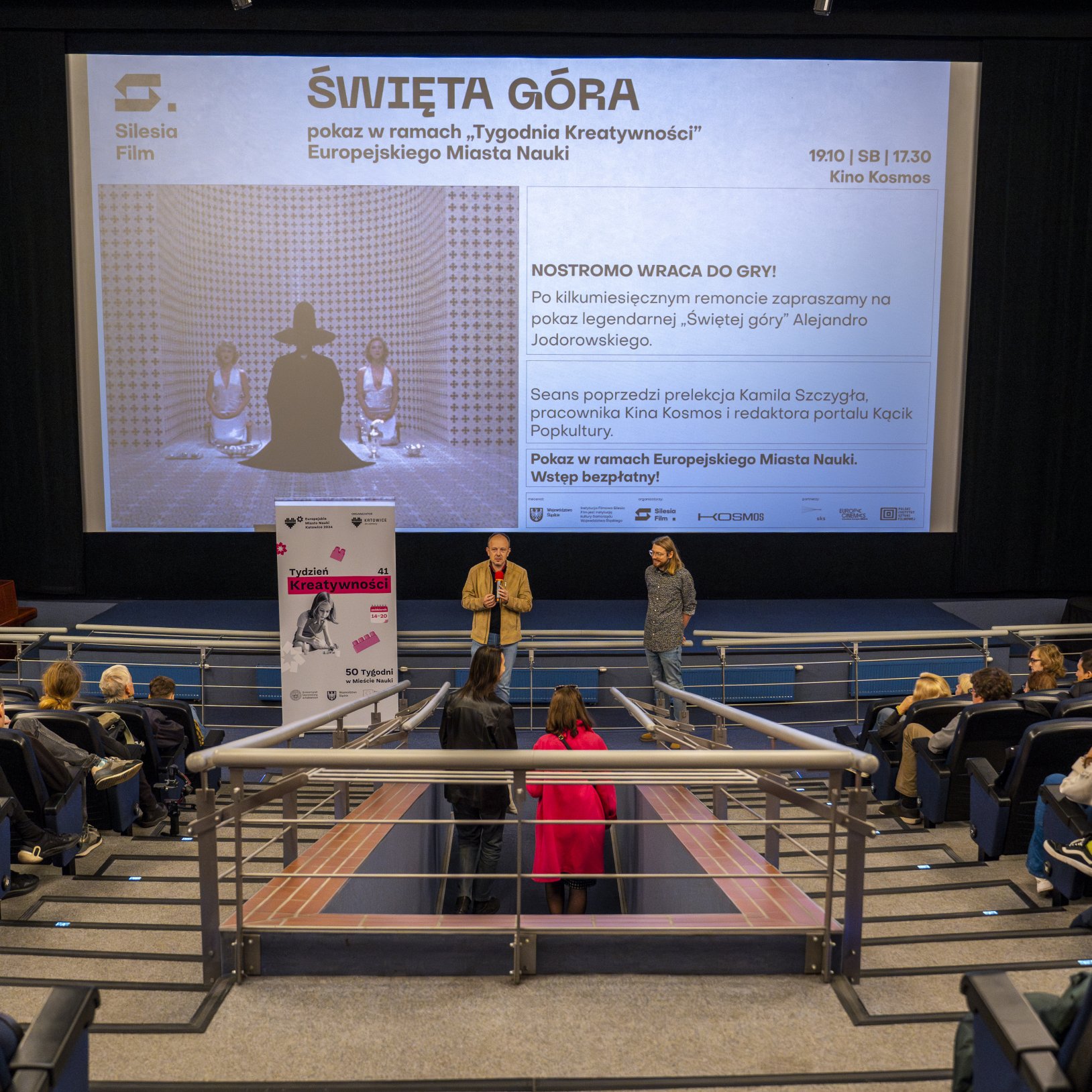 "Sala kinowa z osobą prowadzącą spotkanie przed ekranem z wyświetlonym plakatem filmu 'Święta Góra'. Na widowni siedzą widzowie, a w centralnej części znajduje się zejście z barierkami."
