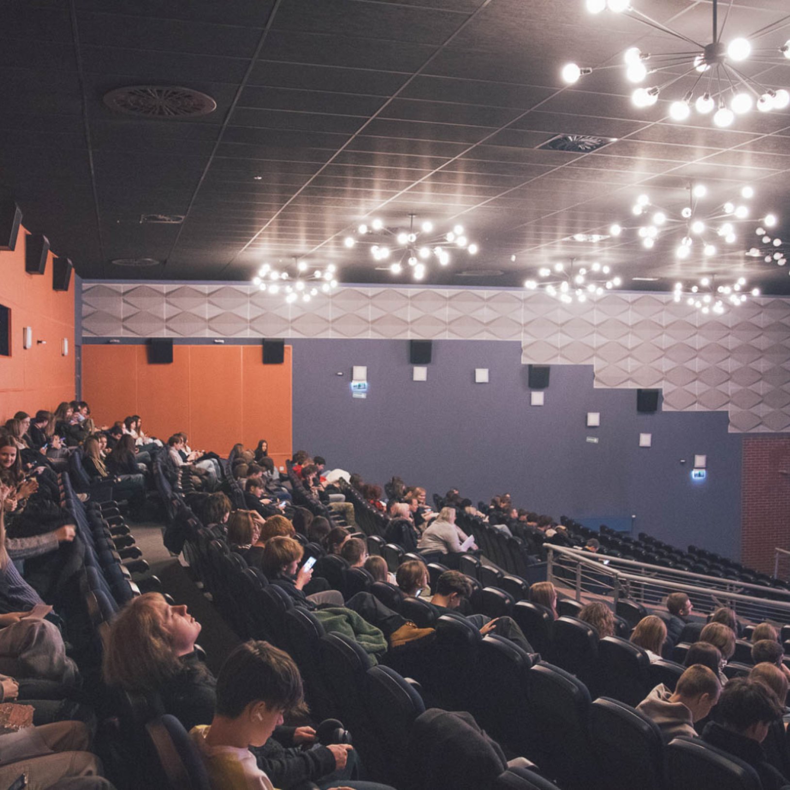"Wnętrze sali kinowej z publicznością siedzącą w ciemnych fotelach. Ściany mają geometryczne panele w odcieniach pomarańczowym i szarym. Sufit ozdobiony nowoczesnym oświetleniem."