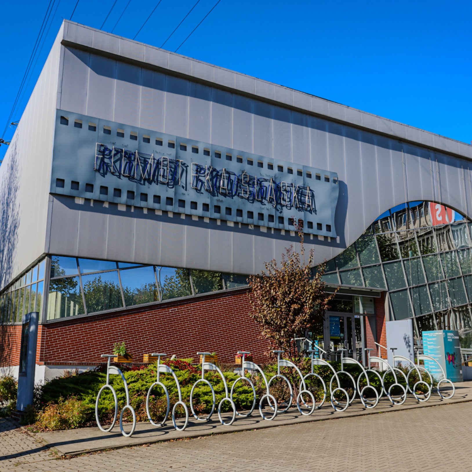 Widok dzienny na Kino Kosmos z zamkniętym neonem. Przed wejściem znajduje się rząd stojaków rowerowych w kształcie stylizowanych rowerów oraz niska roślinność."