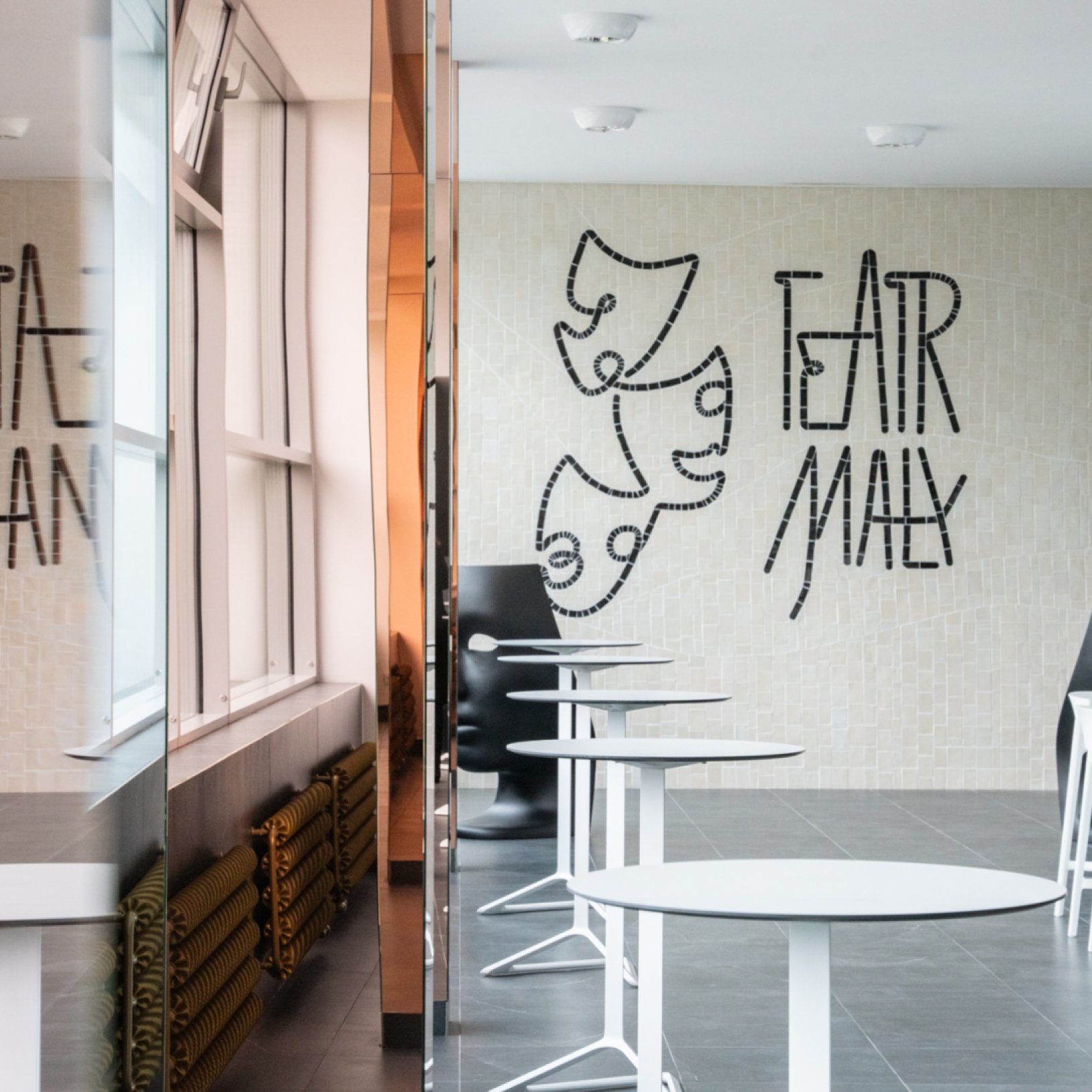 Nowoczesne wnętrze foyer Teatru Małego w Tychach z mozaiką przedstawiającą maski teatralne i napis Teatr Mały na ścianie. W przestrzeni ustawione są białe stoliki i designerskie krzesła.