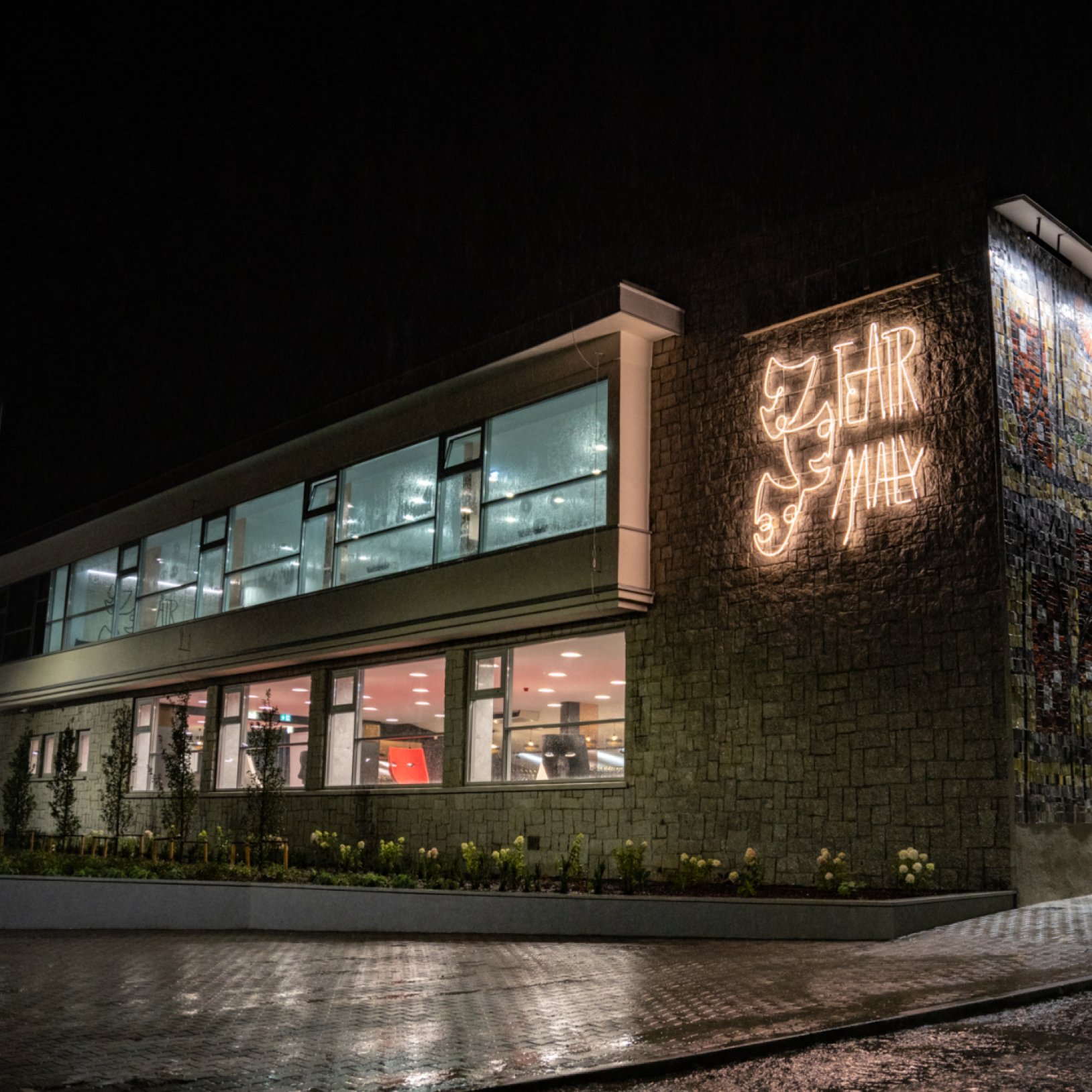 Wieczorny widok na Teatr Mały w Tychach z podświetlonym neonem w kształcie masek teatralnych i napisu Teatr Mały na kamiennej elewacji. Przez okna widać jasno oświetlone wnętrze budynku.