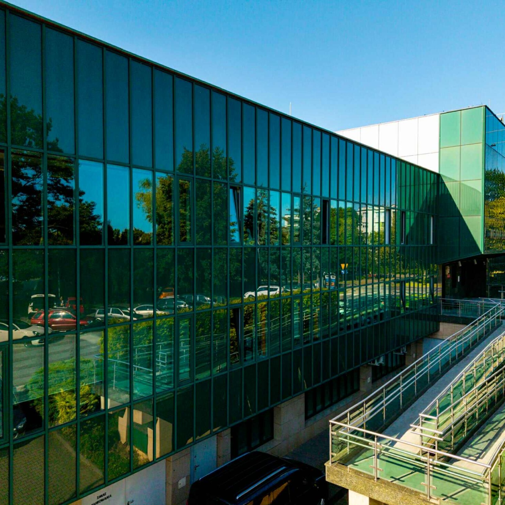 "Boczny widok na przeszkloną elewację Miejskiej Biblioteki Publicznej. W szklanych panelach odbijają się drzewa, ulica i zaparkowane samochody. Wzdłuż budynku biegnie rampa z poręczami."