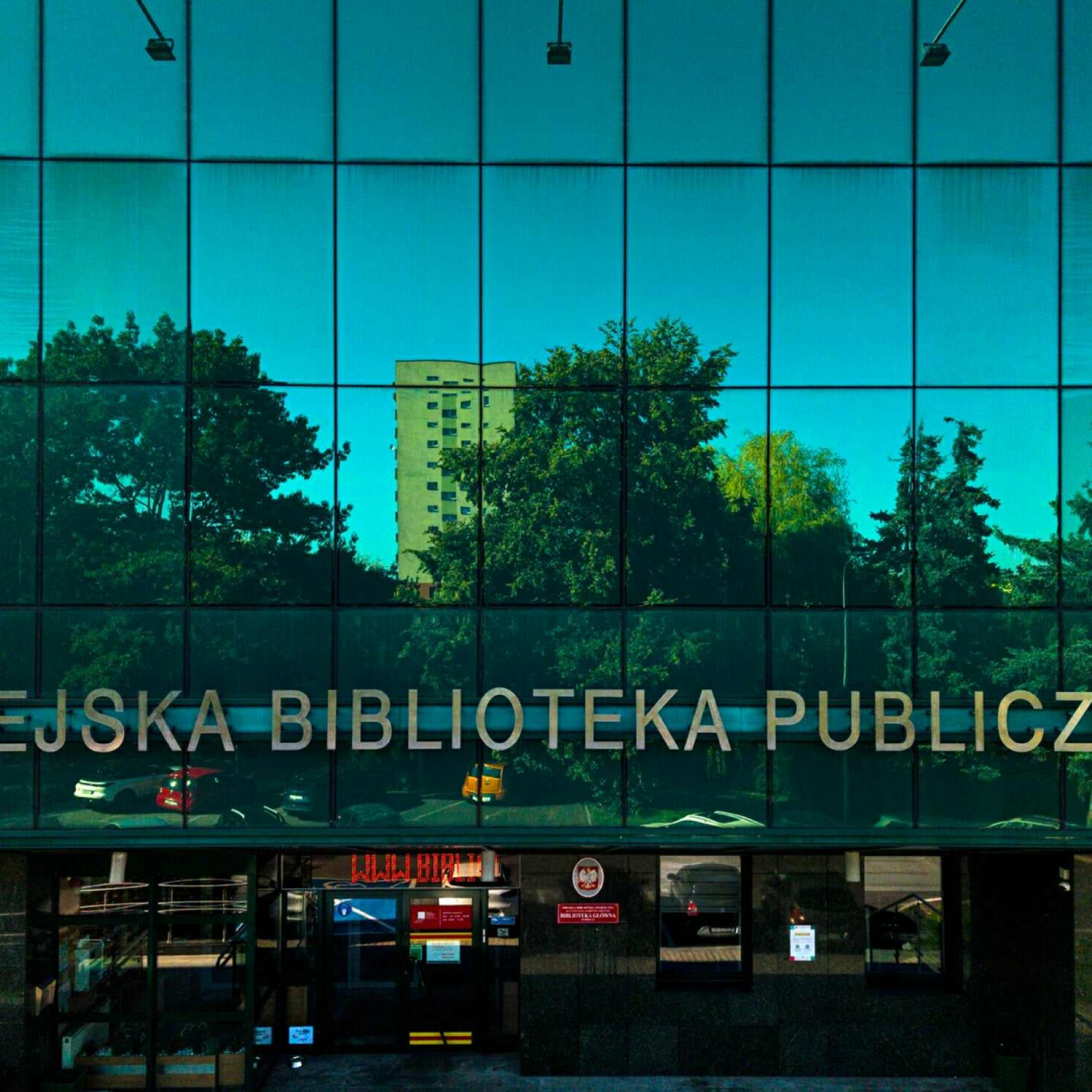 "Widok frontowy nowoczesnego budynku Miejskiej Biblioteki Publicznej z fasadą w całości pokrytą lustrzanym szkłem. W szklanej elewacji odbijają się drzewa oraz wysoki blok mieszkalny."