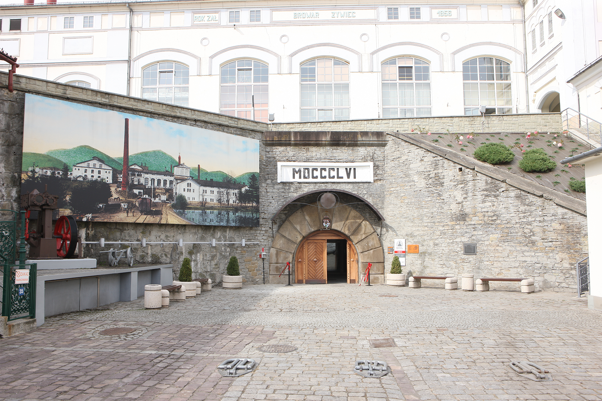 "Wejście do Muzeum Browaru Żywiec, umiejscowione w zabytkowym kamiennym budynku z datą „MDCCCLVI” (1856) nad portalem. Po lewej stronie znajduje się mural przedstawiający dawny wygląd browaru.