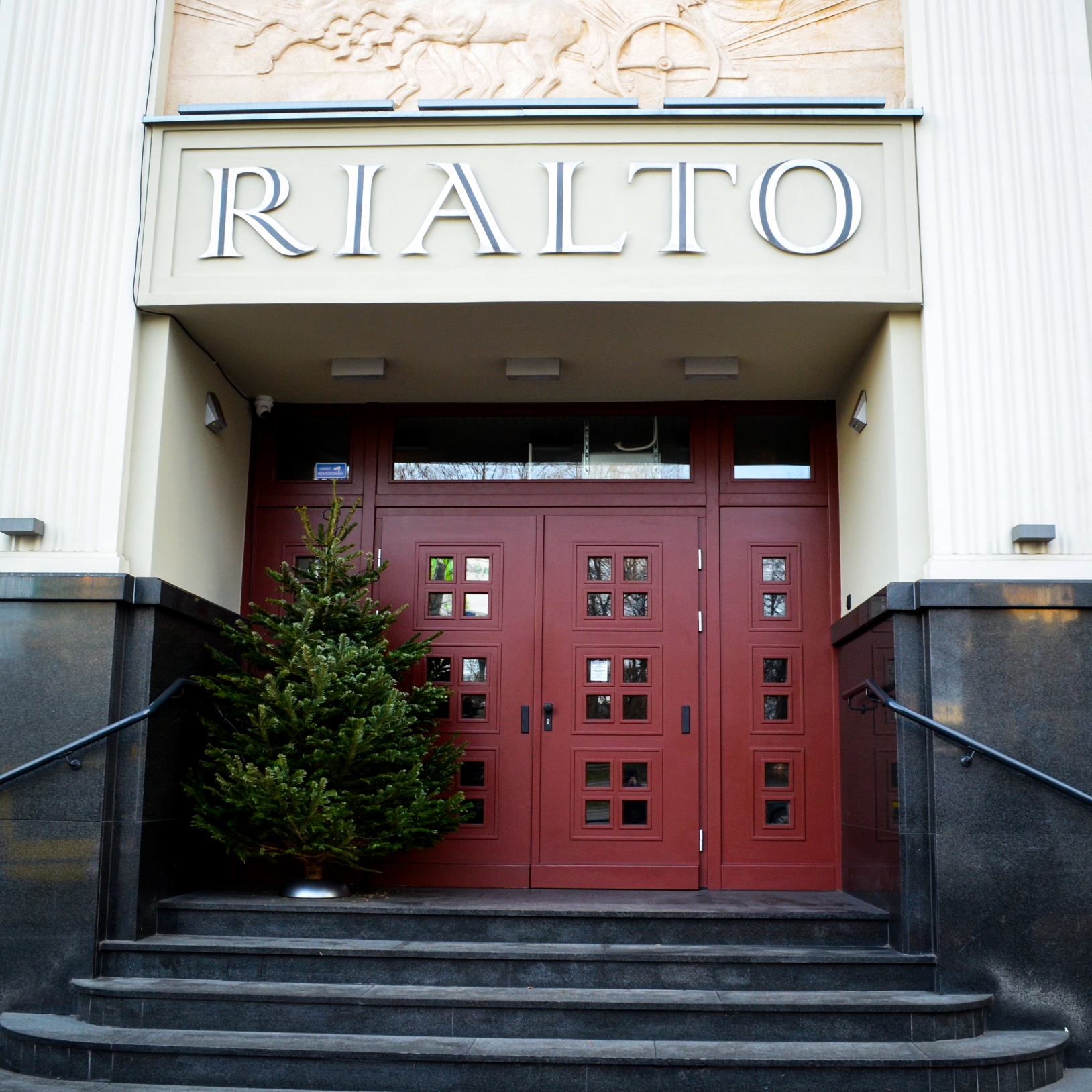"Wejście do Kinoteatru Rialto z czerwonymi drzwiami i świąteczną choinką ustawioną po lewej stronie schodów. Nad wejściem widoczny jest duży napis 'RIALTO' oraz płaskorzeźba z motywem rydwanu. Budynek utrzymany w stylu modernistycznym z jasną elewacją."