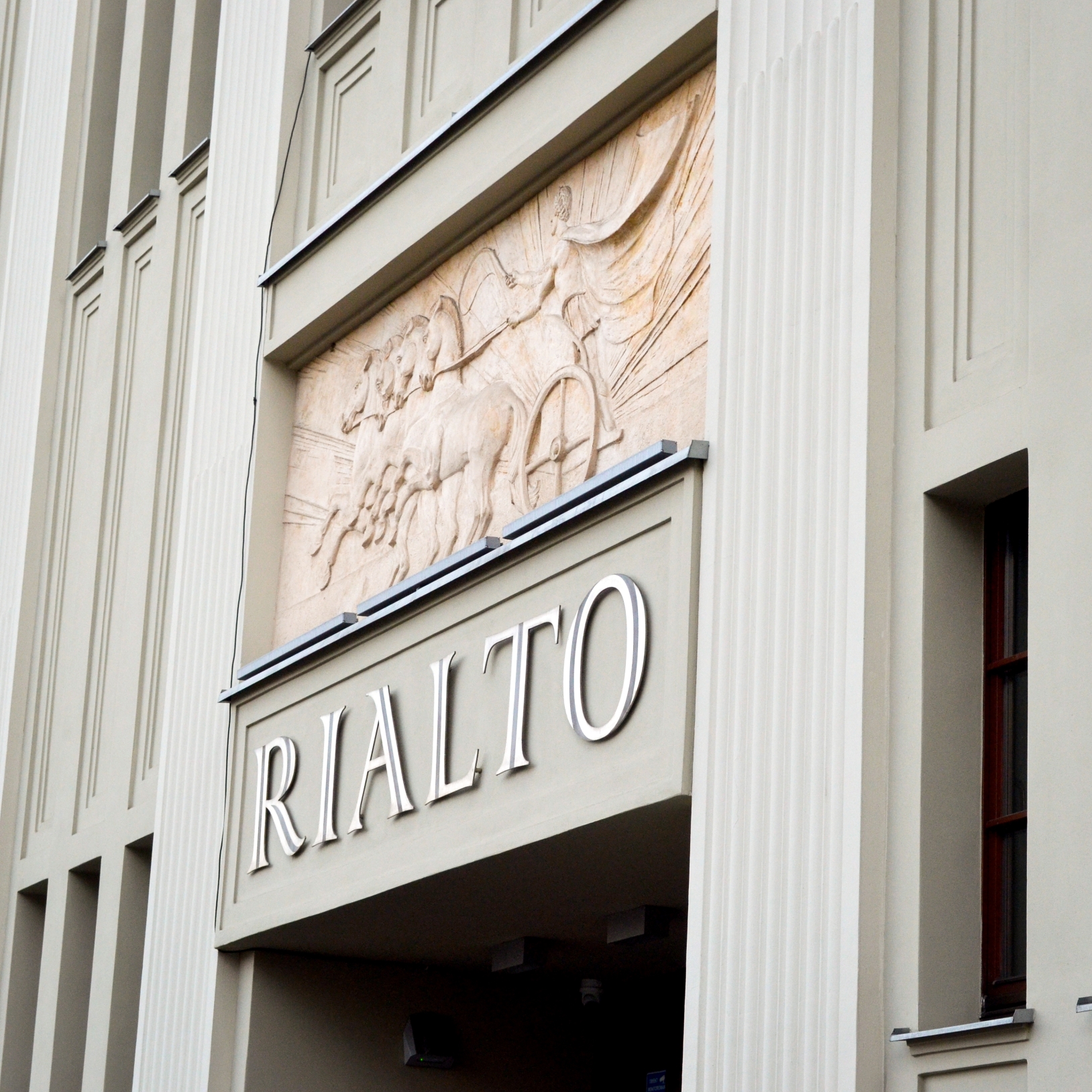 "Zbliżenie na elewację Kinoteatru Rialto z widocznym napisem 'RIALTO' oraz płaskorzeźbą przedstawiającą rydwan zaprzężony w konie. Budynek utrzymany jest w stylu modernistycznym z pionowymi podziałami i jasnym kolorem elewacji."