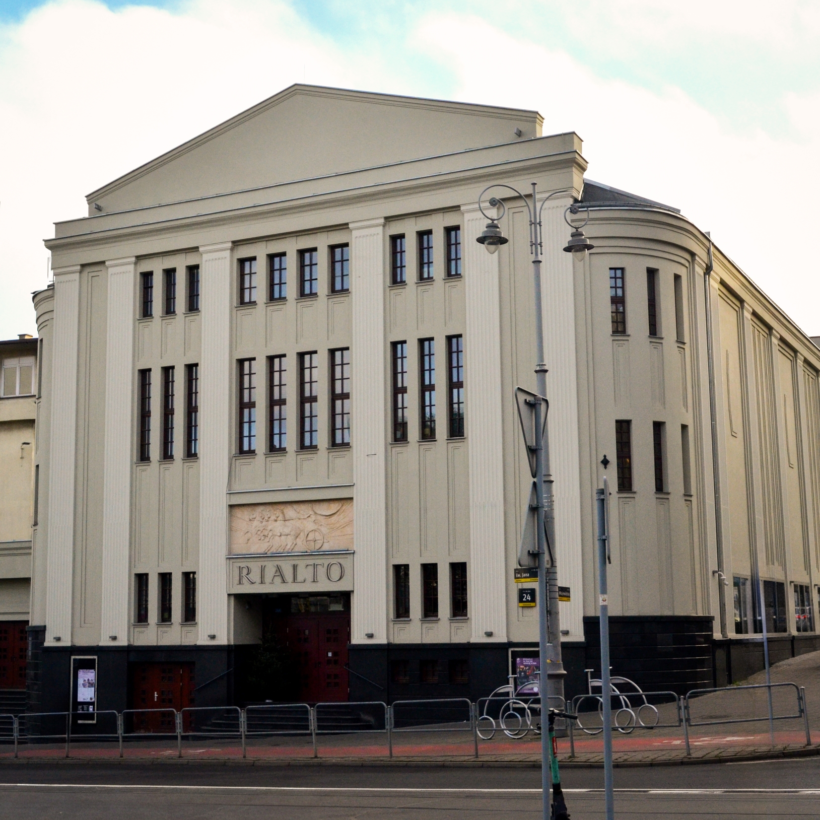 "Widok zewnętrzny na cały budynek Kinoteatru Rialto. Elewacja modernistyczna w jasnym kolorze z dużą ilością okien oraz napisem 'RIALTO' nad głównym wejściem. Przed budynkiem znajduje się przystanek komunikacji miejskiej i stojaki na rowery."