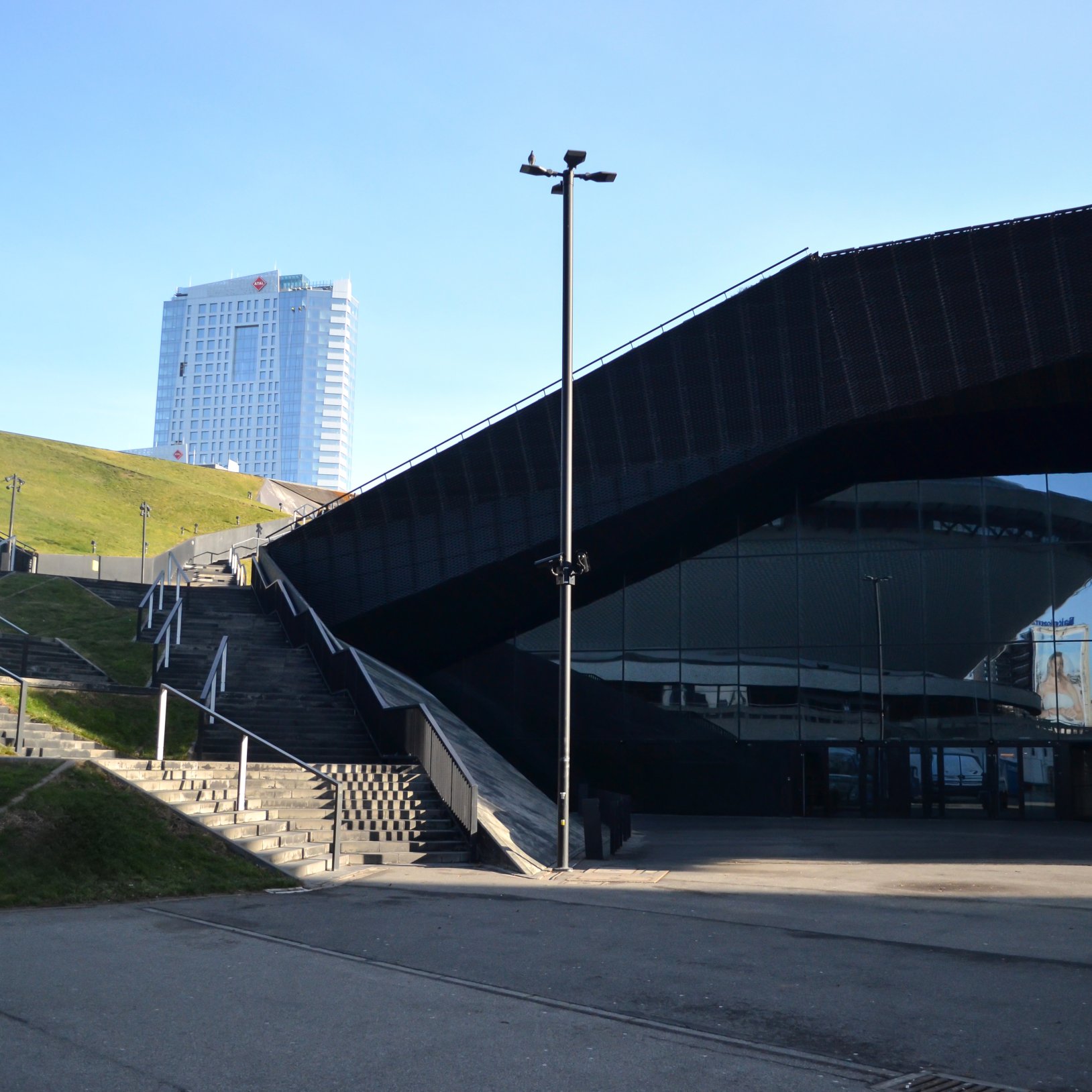 "Widok na schody prowadzące do nowoczesnego budynku z ciemną elewacją i przeszklonym wejściem. W odbiciu szyb widoczna jest charakterystyczna bryła hali Spodek. W tle, na wzgórzu, znajduje się wysoki biurowiec z logo hotelu. Teren wokół jest pokryty trawą