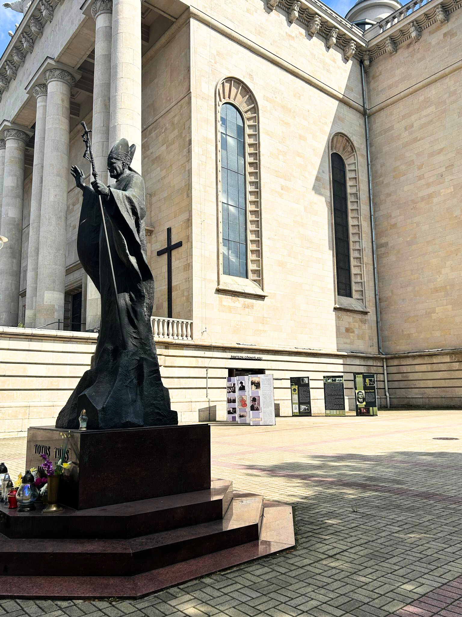 "Pomnik papieża Jana Pawła II ustawiony przed monumentalną fasadą katedry z jasnego kamienia. Wokół cokołu widoczne są znicze i kwiaty. W tle widać wystawę plenerową z planszami informacyjnymi oraz wysokie okna katedry i kolumny."