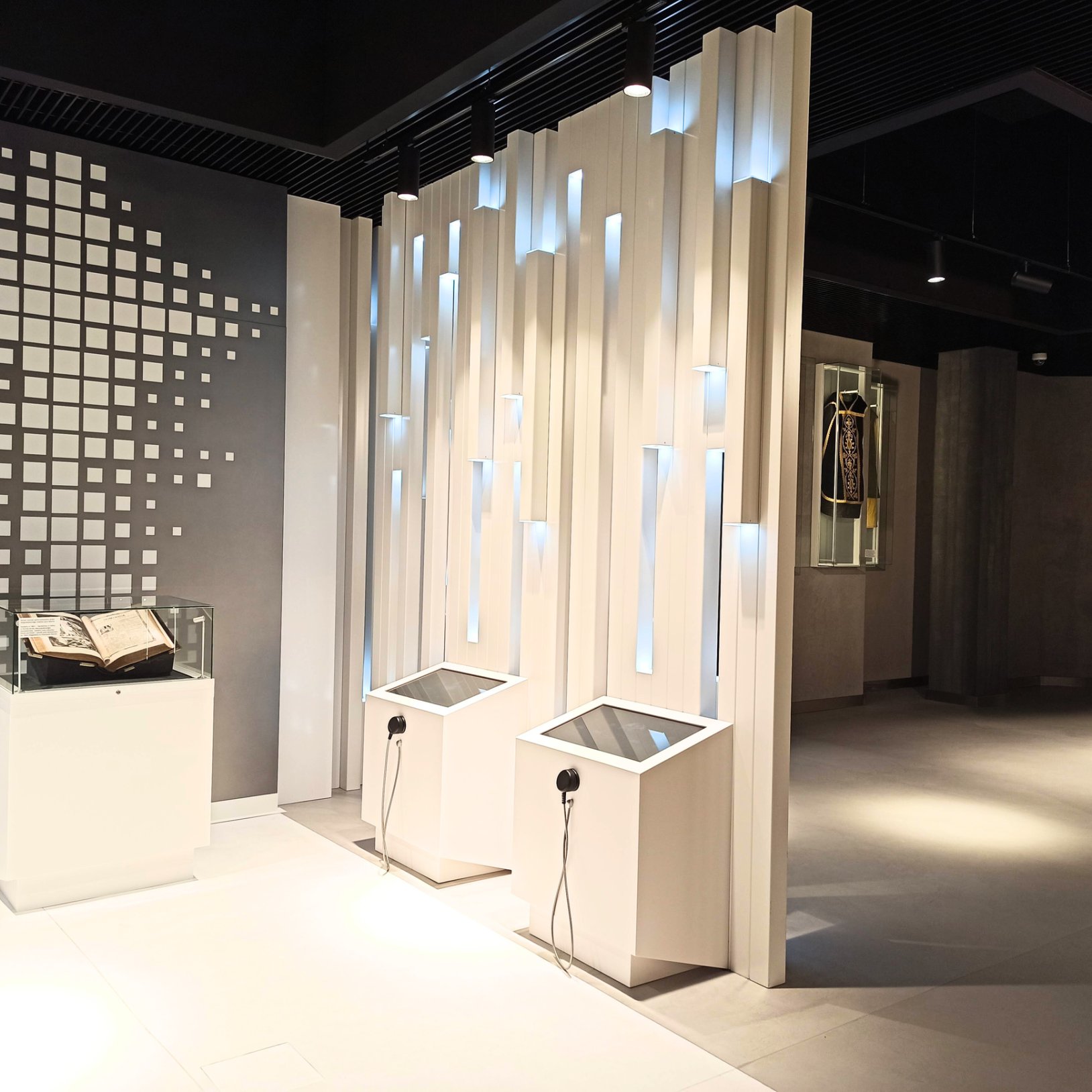 "Wnętrze nowoczesnej przestrzeni muzealnej z interaktywnymi stanowiskami multimedialnymi i podświetlaną dekoracją ścienną z pionowych, białych paneli. Po lewej stronie znajduje się zamknięta w gablocie książka.